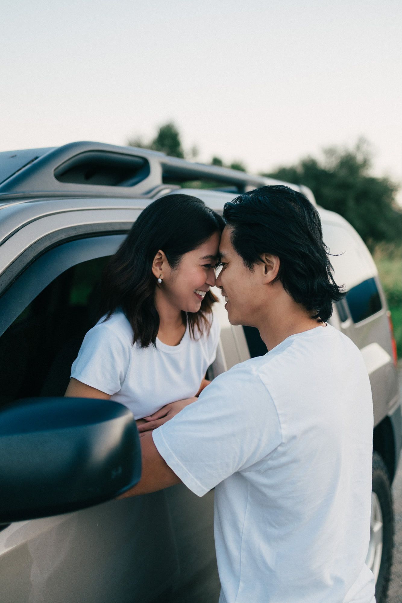 Smiling couple touching foreheads beside a silver SUV during an outdoor engagement photo session at sunset.