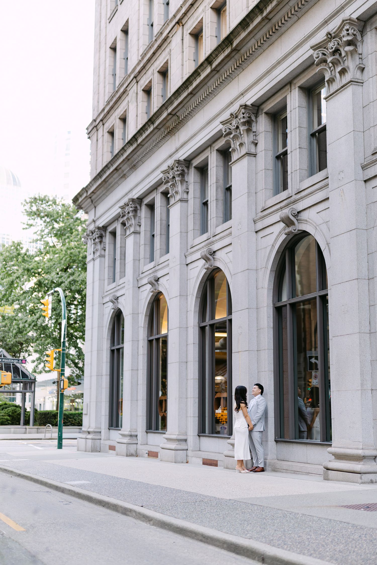 Couple stands close together beside tall arched windows of a historic stone building on a quiet city street.