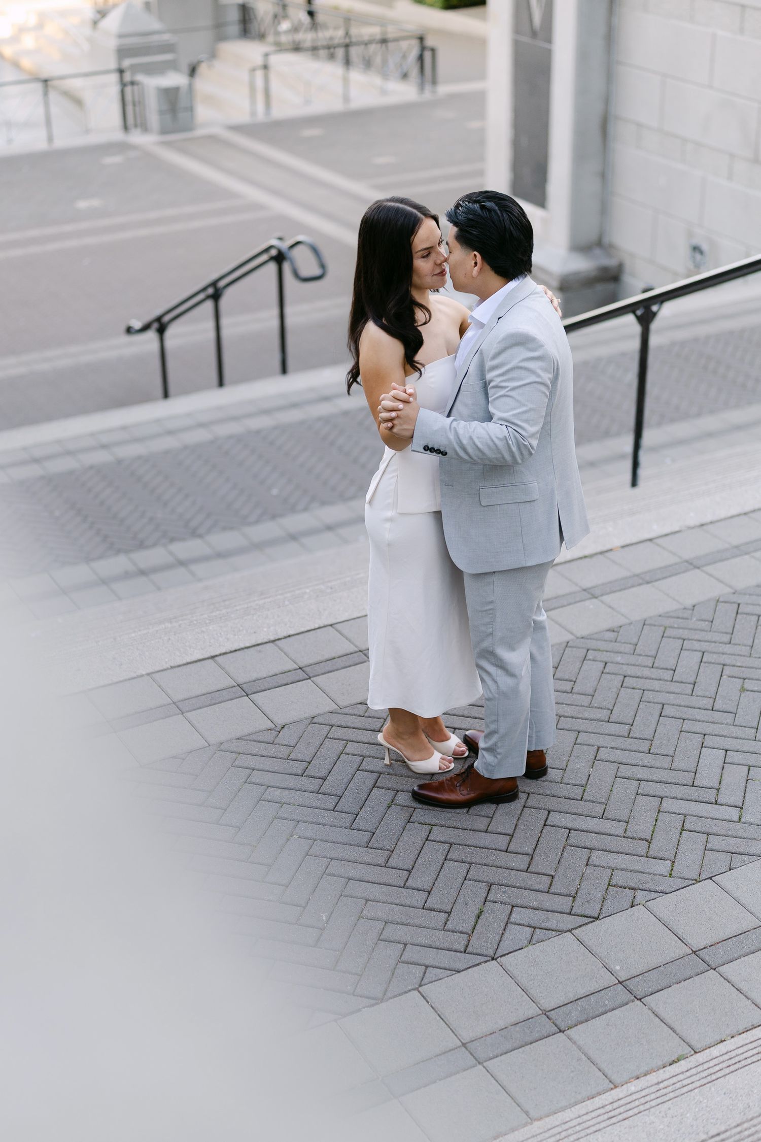 Couple in a gray suit and white dress embrace on brick-paved steps in an intimate, romantic city engagement photo.