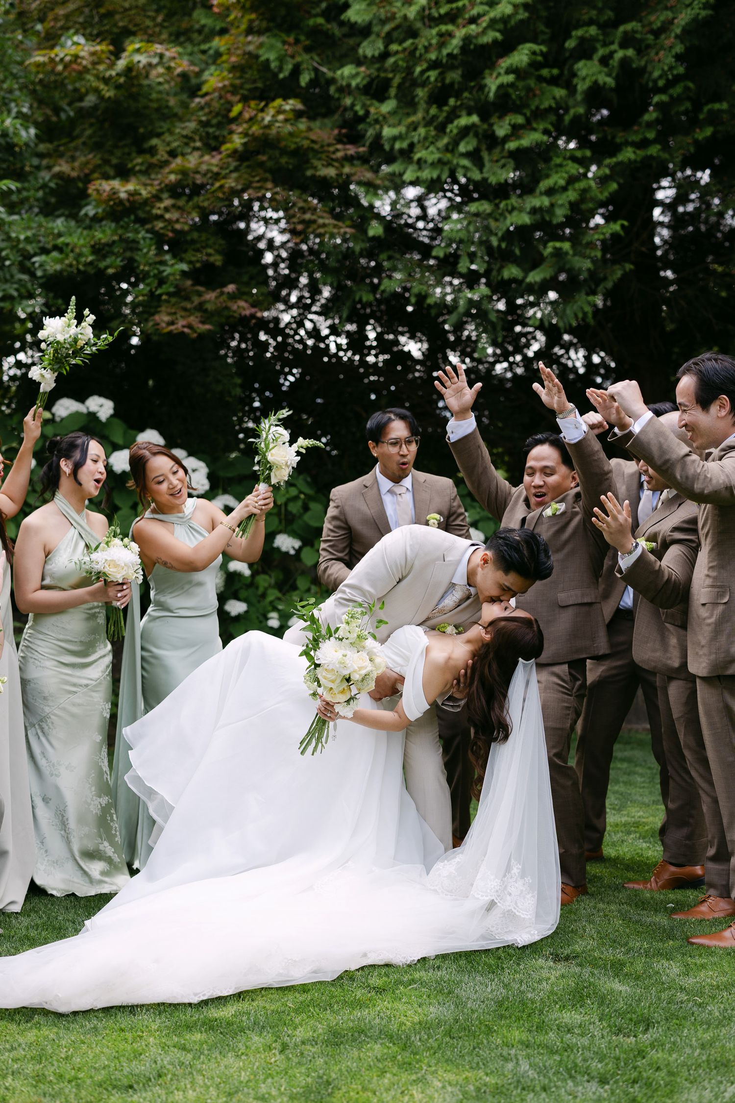 Wedding party celebrates as groom dips bride on lush green lawn with bridesmaids in sage gowns.
