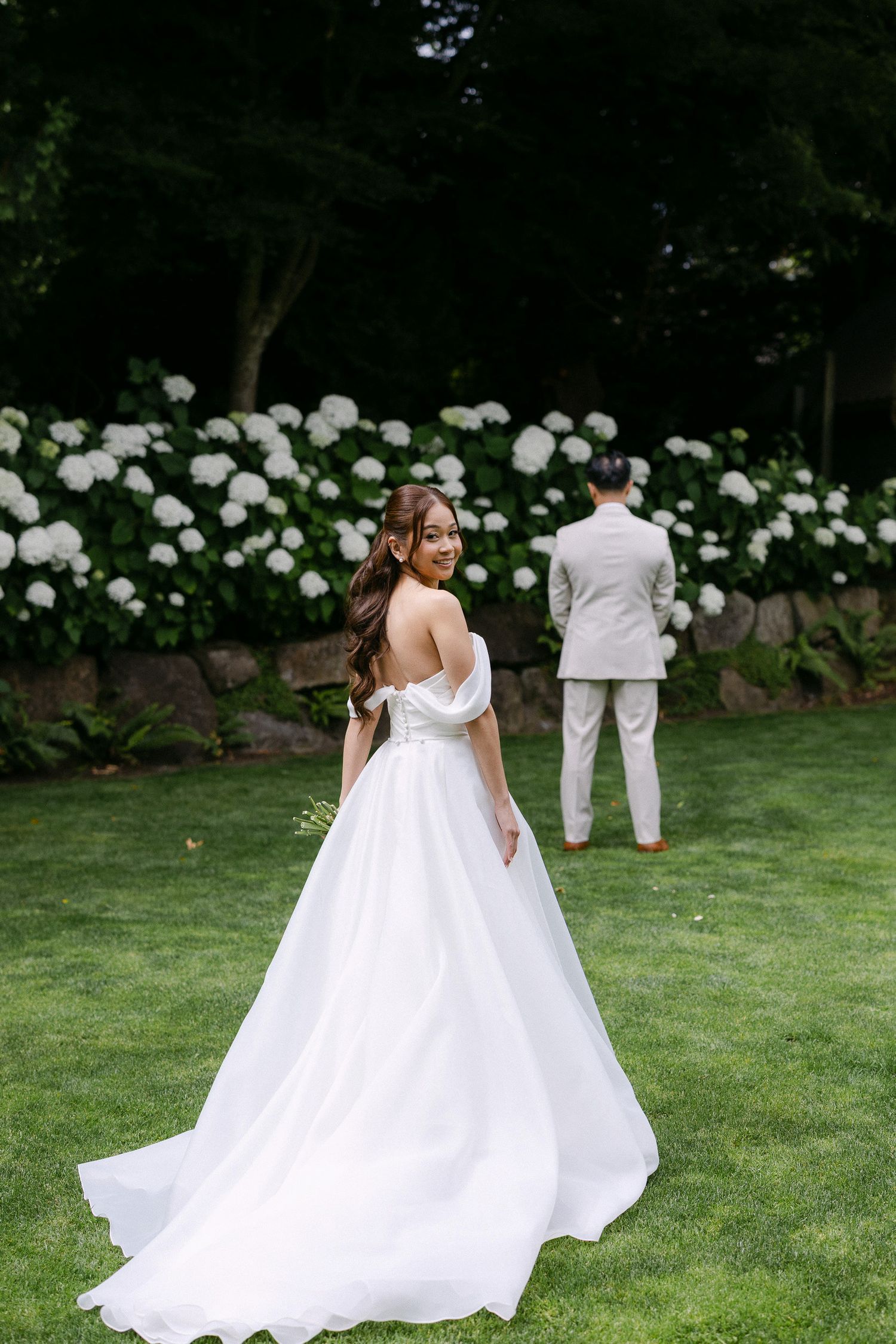 Bride in white off-shoulder ballgown looks back smiling, groom in tan suit stands before white hydrangeas on green lawn.