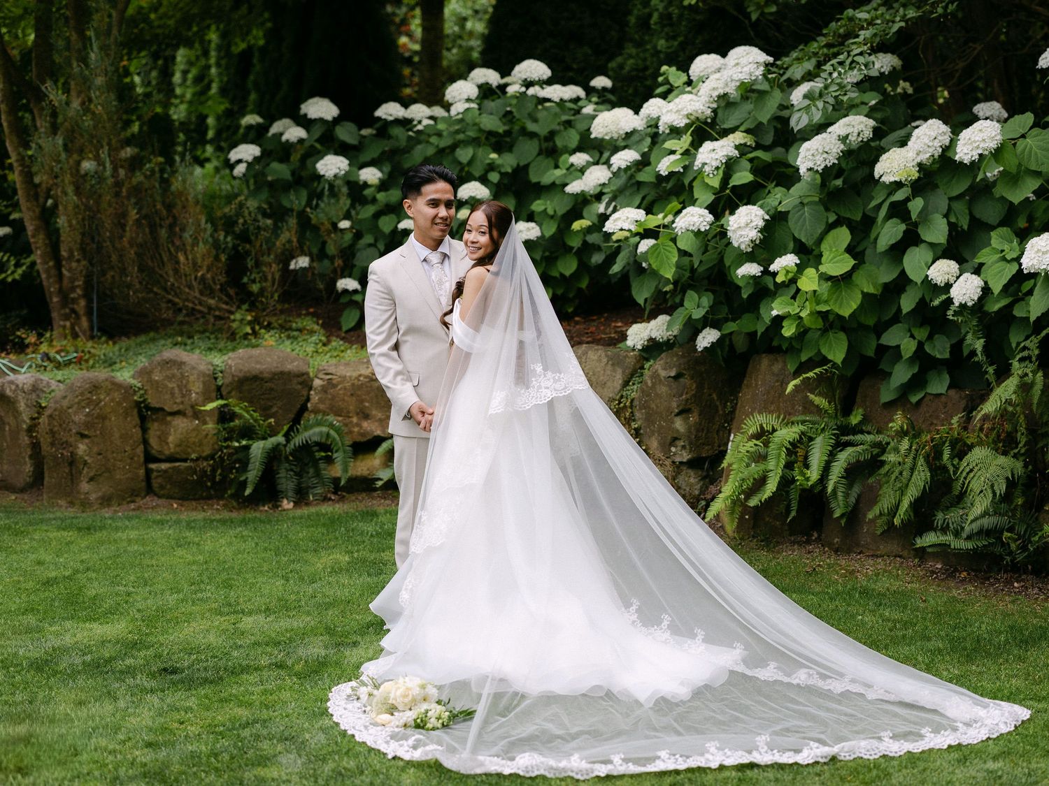 Bride and groom posing in lush garden with white hydrangeas, bride wearing long veil and white gown, groom in beige suit.