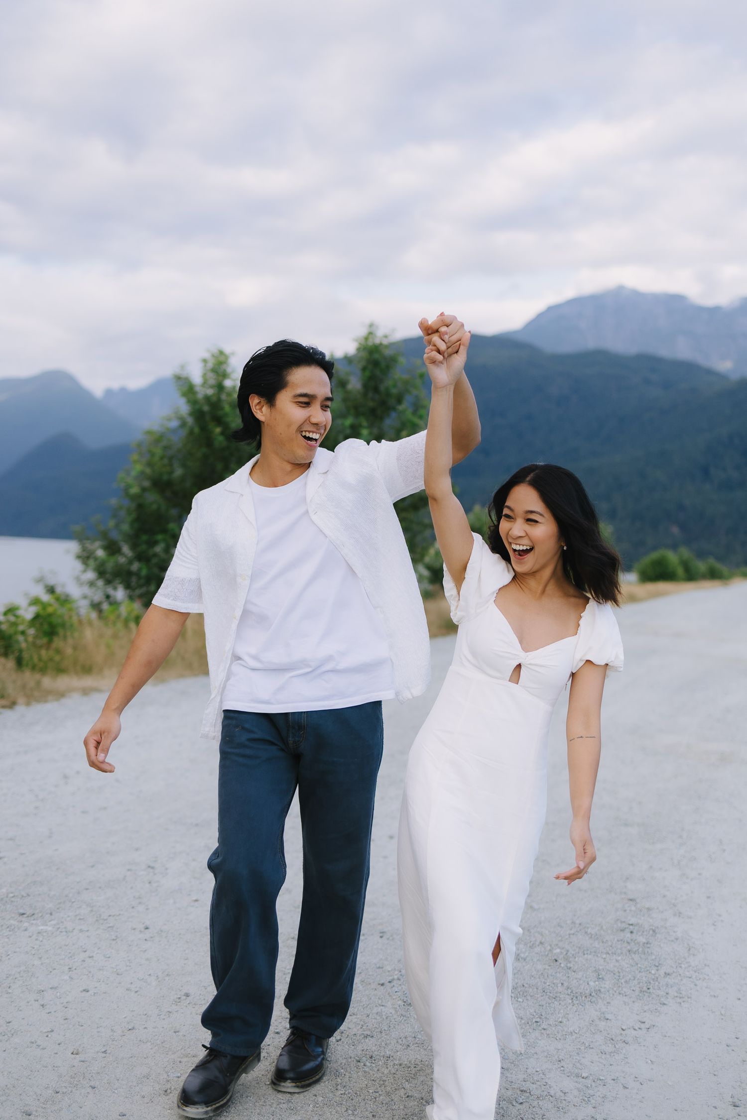 A couple shares a joyful dance moment on a gravel path with mountains and greenery in the background.