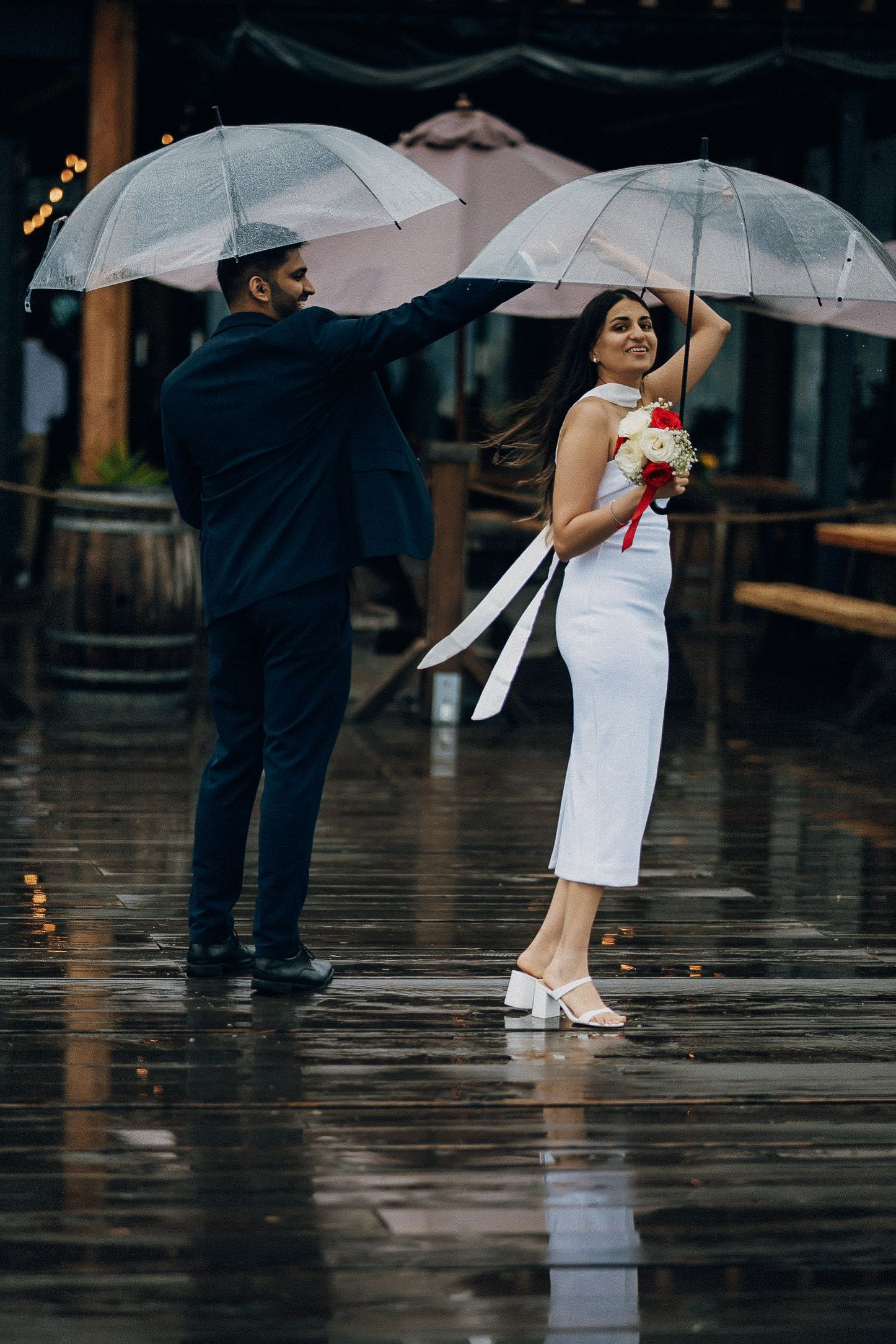 A couple holding white umbrellas stands close together on wet pavement during a rainy wedding photoshoot.