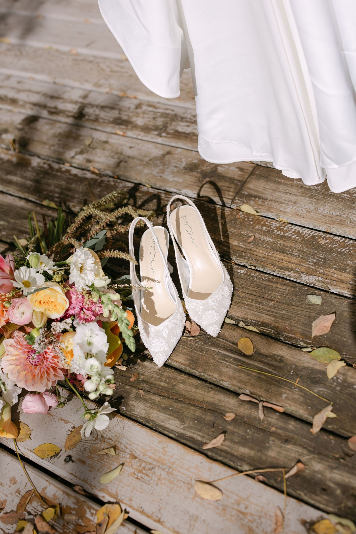 White lace bridal heels beside a pastel wedding bouquet on rustic wooden boards with a flowing white gown above.