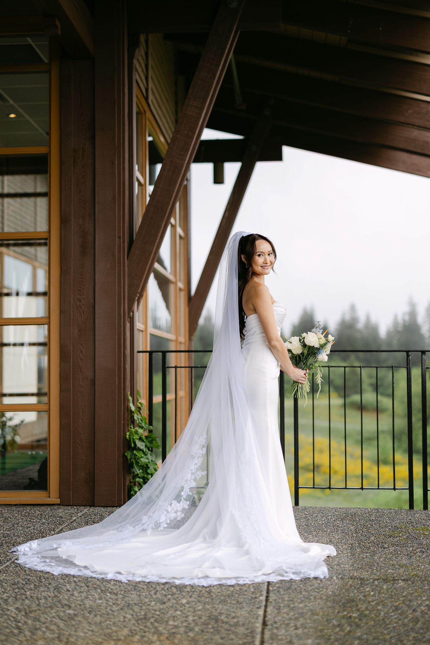 Bride in strapless white gown with long lace cathedral veil holding bouquet on outdoor venue balcony.