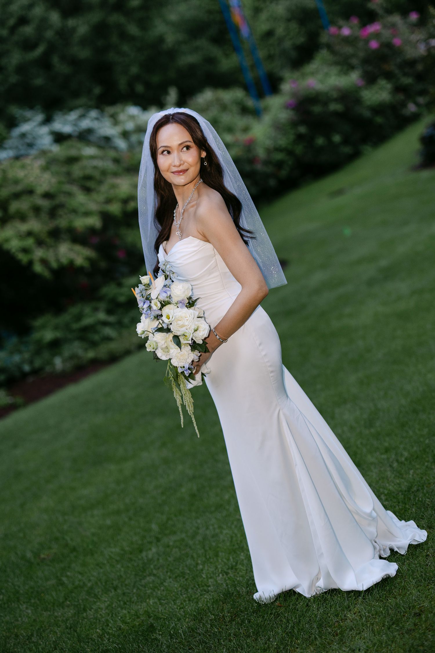 Elegant bride in white strapless mermaid gown and veil holding white bouquet on lush green lawn.