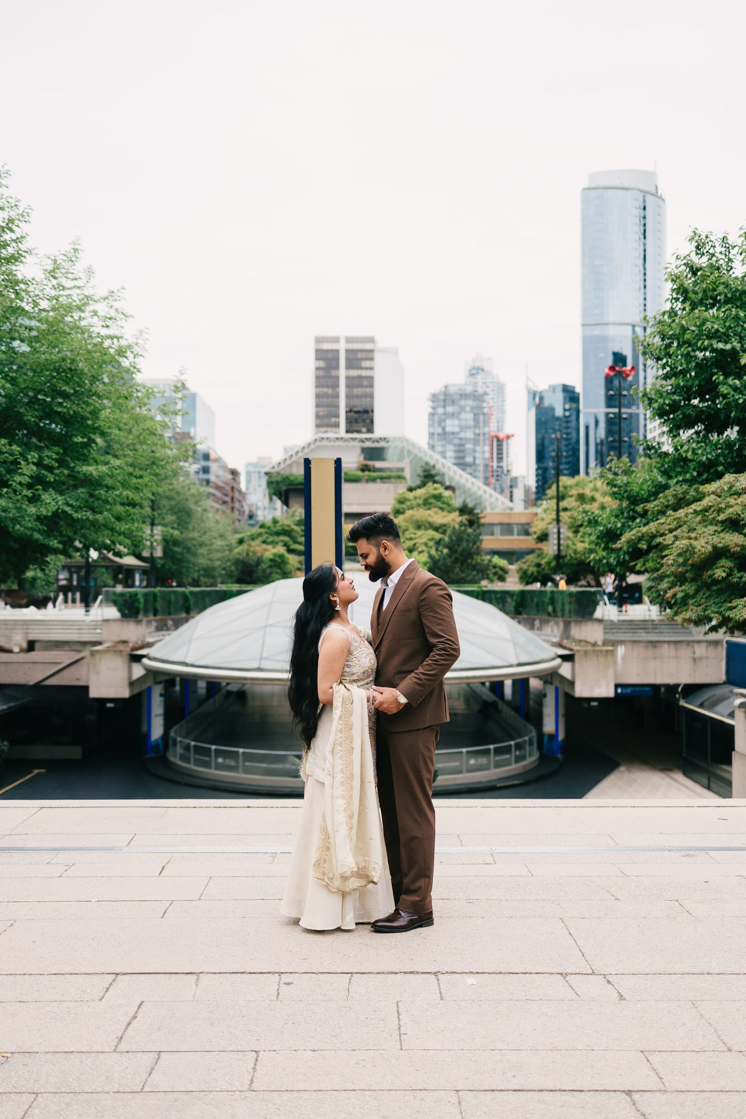 A couple poses romantically on an urban plaza with city skyline and trees in the background.