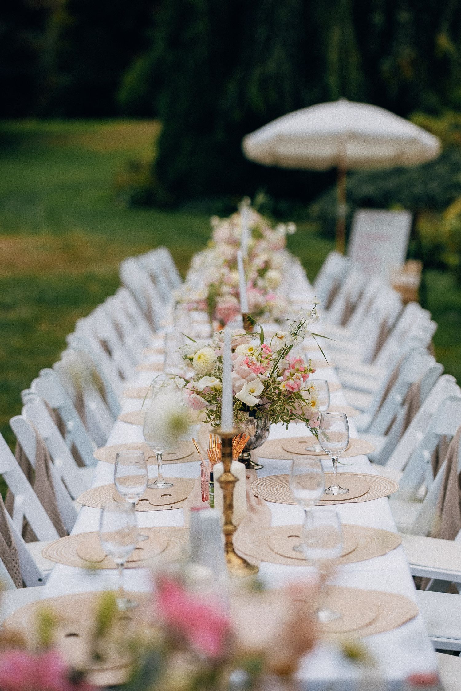 Elegant outdoor wedding table setting with white chairs and pink floral centerpieces under a garden umbrella.