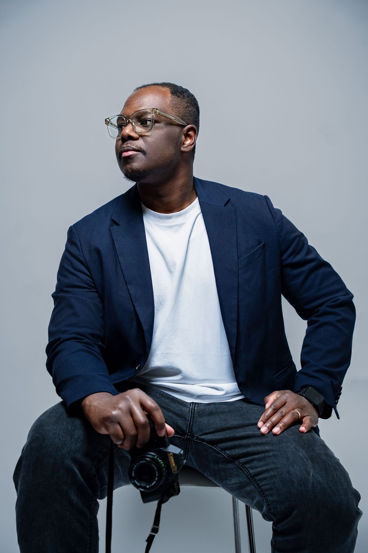 Professional photographer in navy blazer and white t-shirt sits with camera against gray studio backdrop.