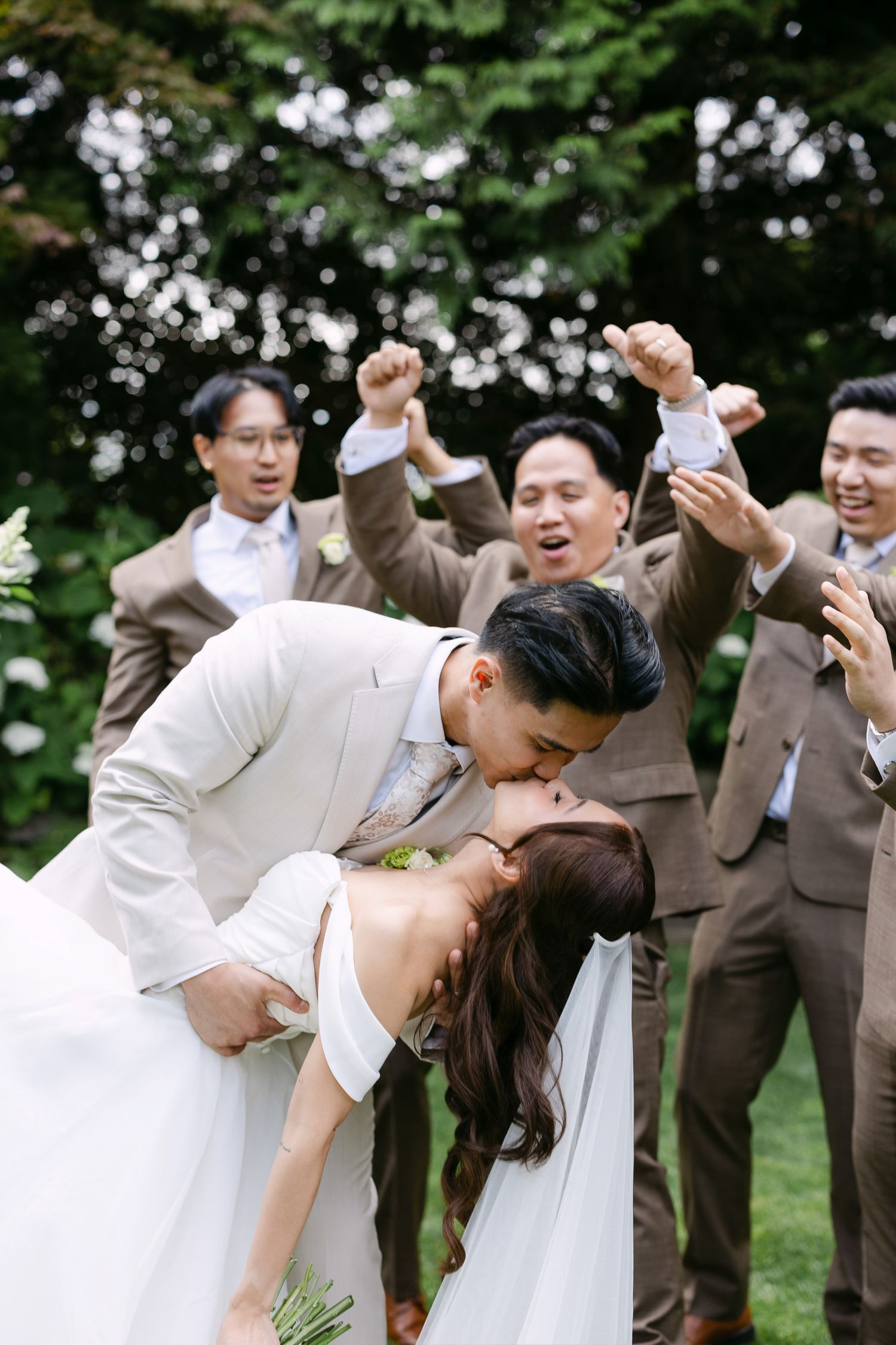Groom dips bride for a kiss while groomsmen in brown suits celebrate joyfully outdoors.