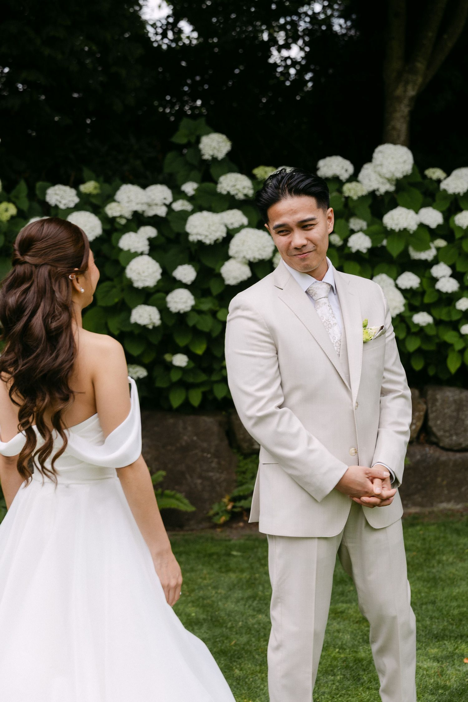 Groom in beige suit smiles at bride in white off-shoulder gown during outdoor wedding ceremony with hydrangeas.