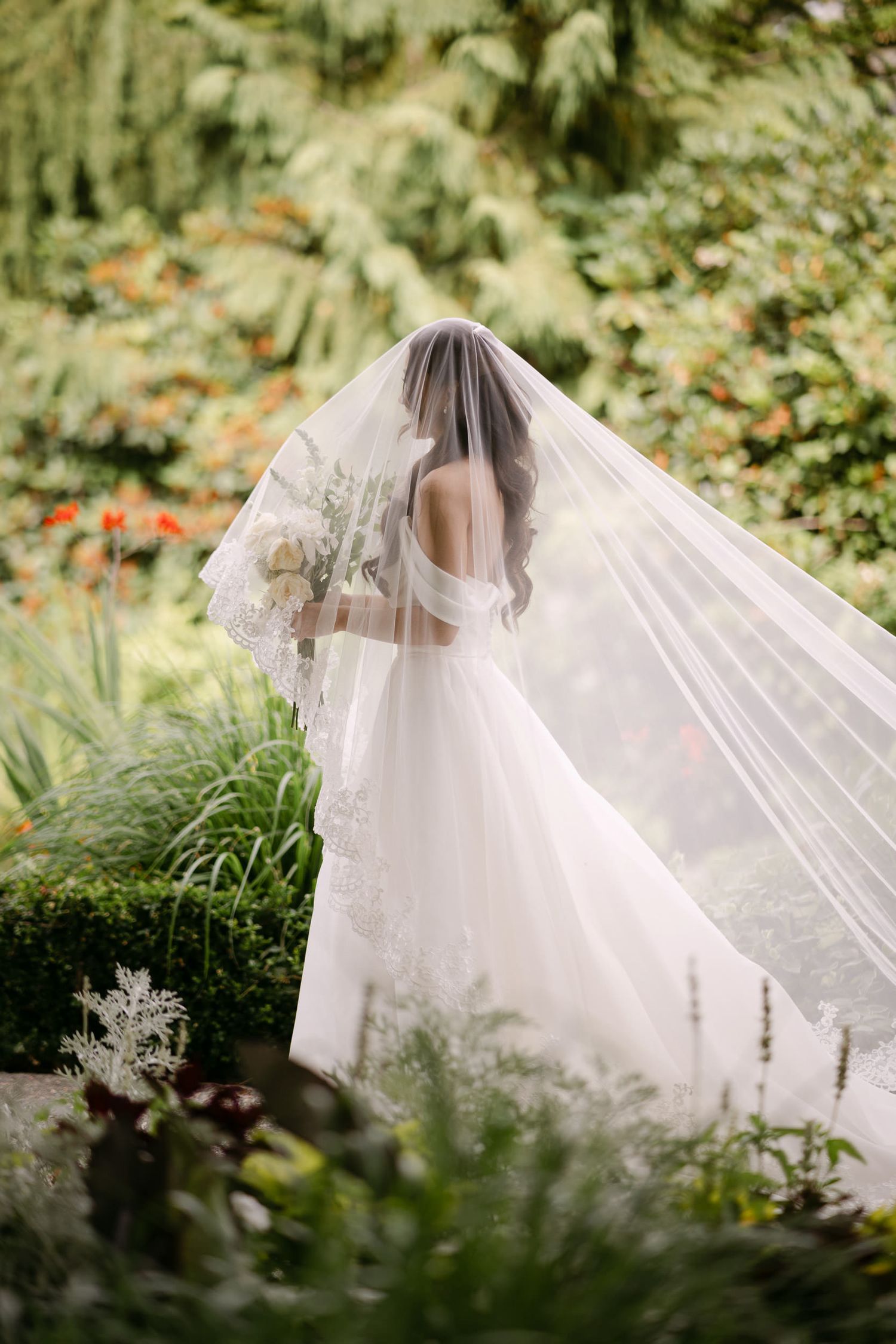 Bride in elegant white wedding gown with flowing veil holding bouquet in lush garden setting.