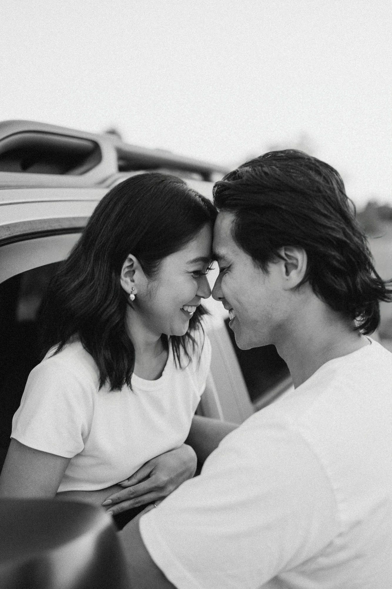 Black and white photo of a smiling couple in white t-shirts touching foreheads beside a vehicle in a tender, romantic moment.