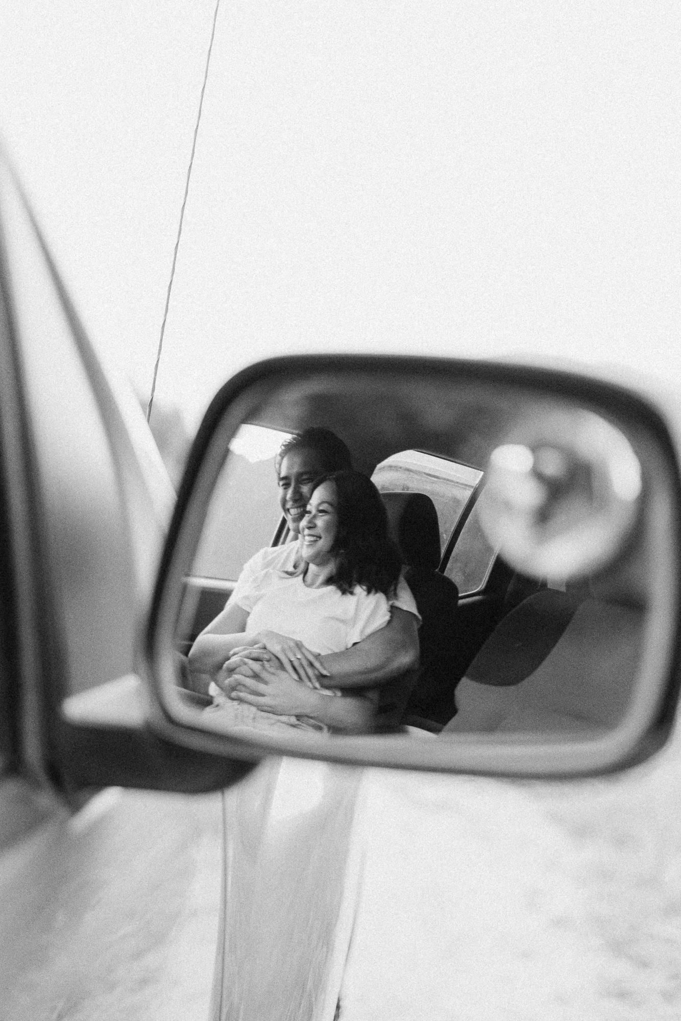 Black and white reflection in a car side mirror showing a smiling couple embracing inside the vehicle.