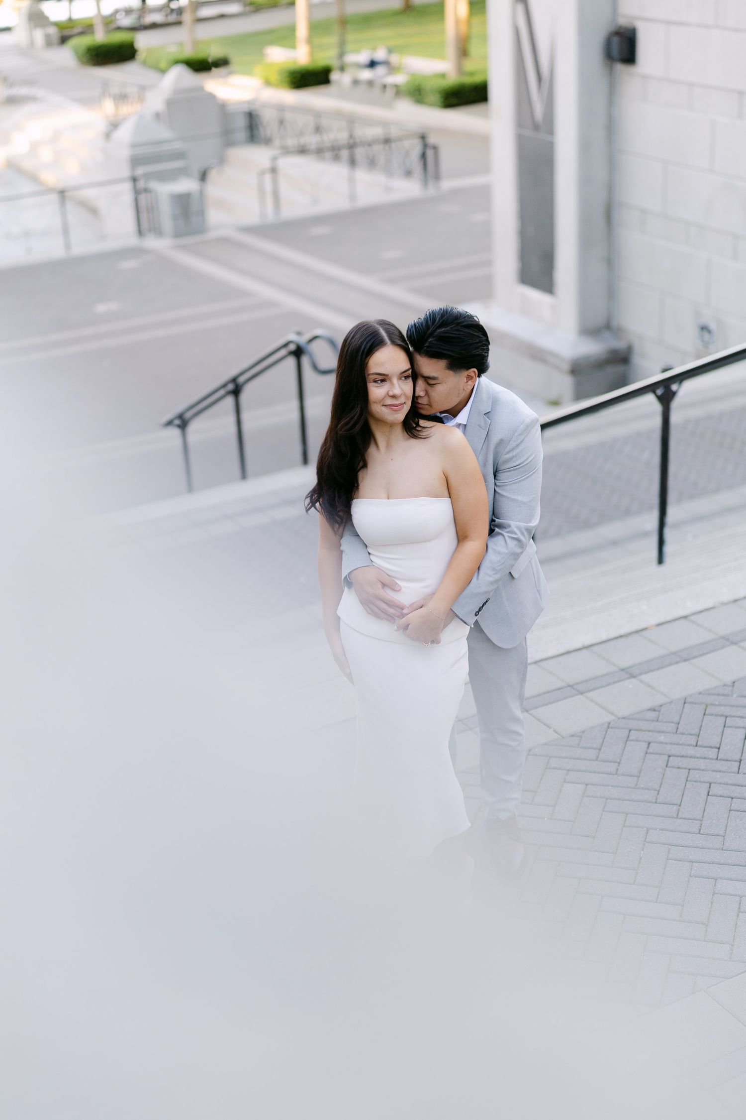 A man in a light gray suit embraces a woman in a white strapless dress from behind on outdoor stone steps with metal railings.
