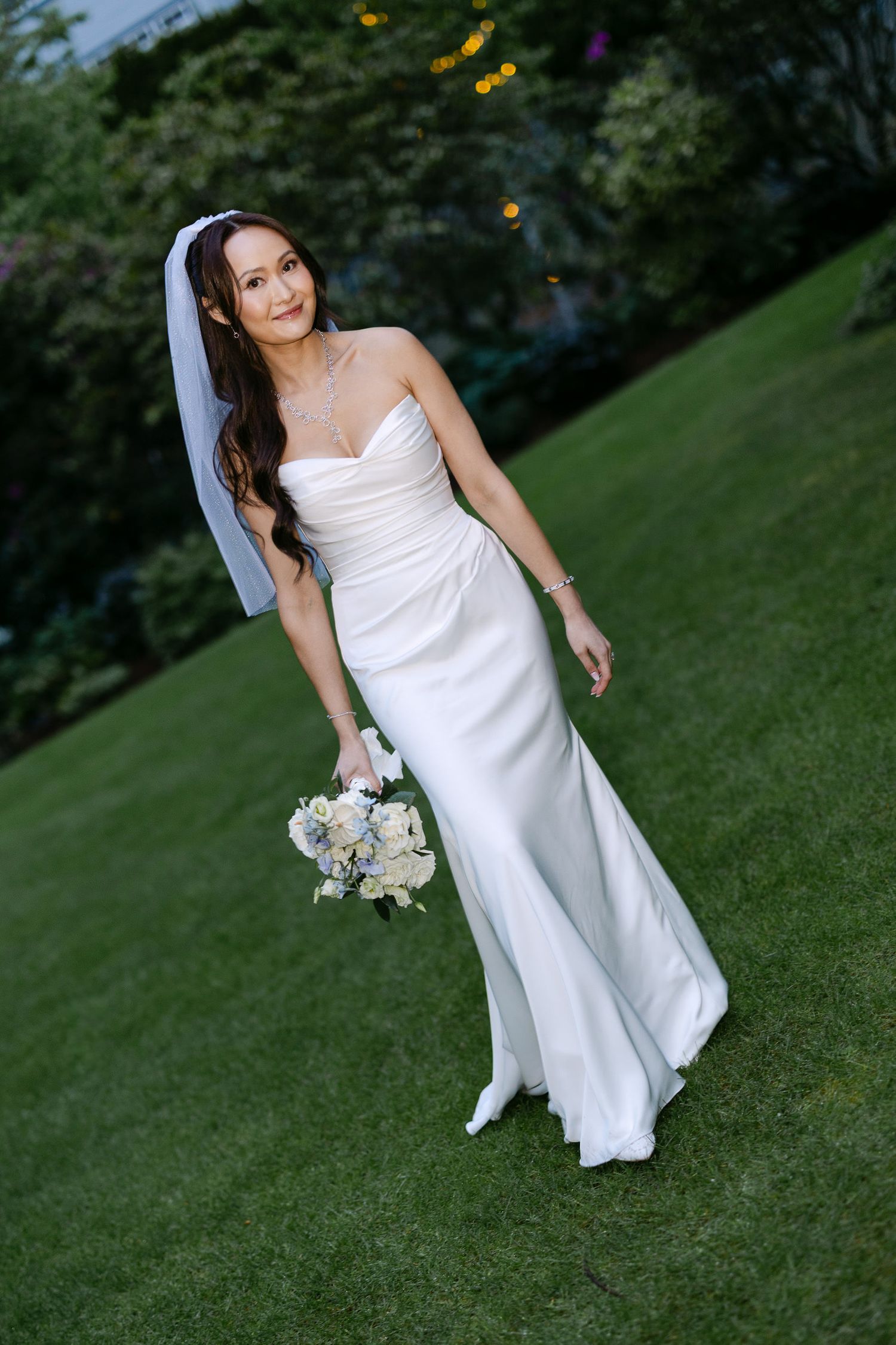 Bride in strapless white gown holds bouquet while walking on lush green lawn at dusk.