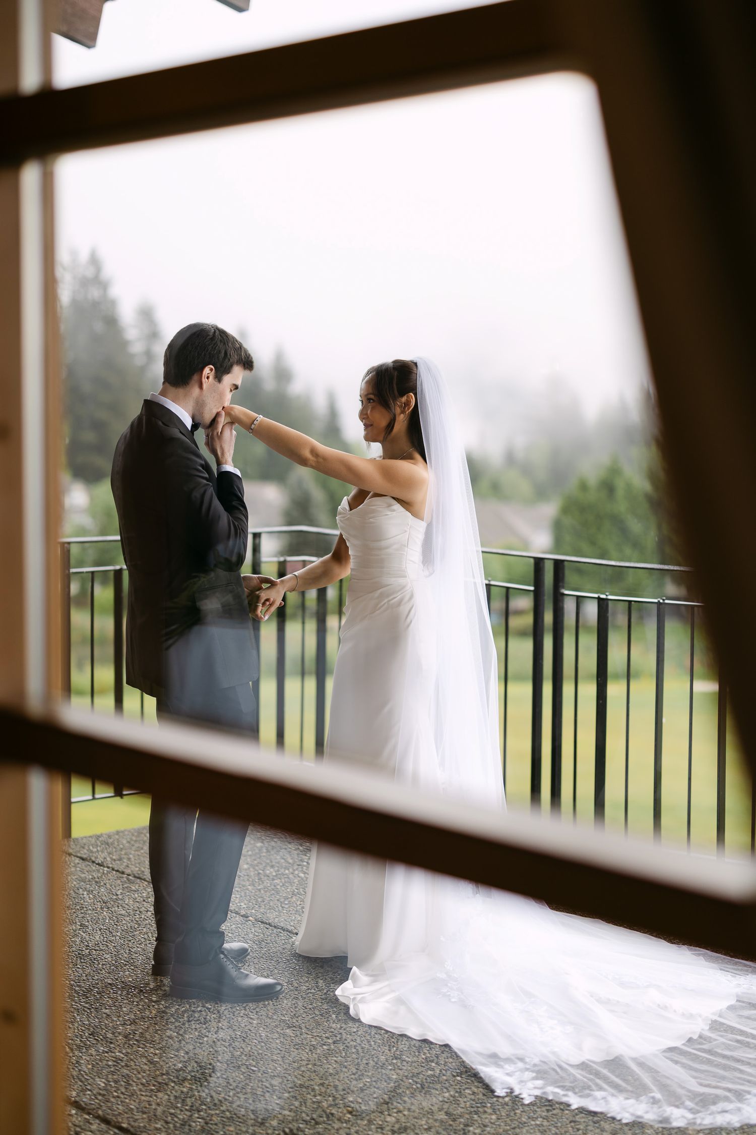 Groom kisses bride's hand on rainy outdoor balcony, viewed through window frame, bride in white gown with veil.