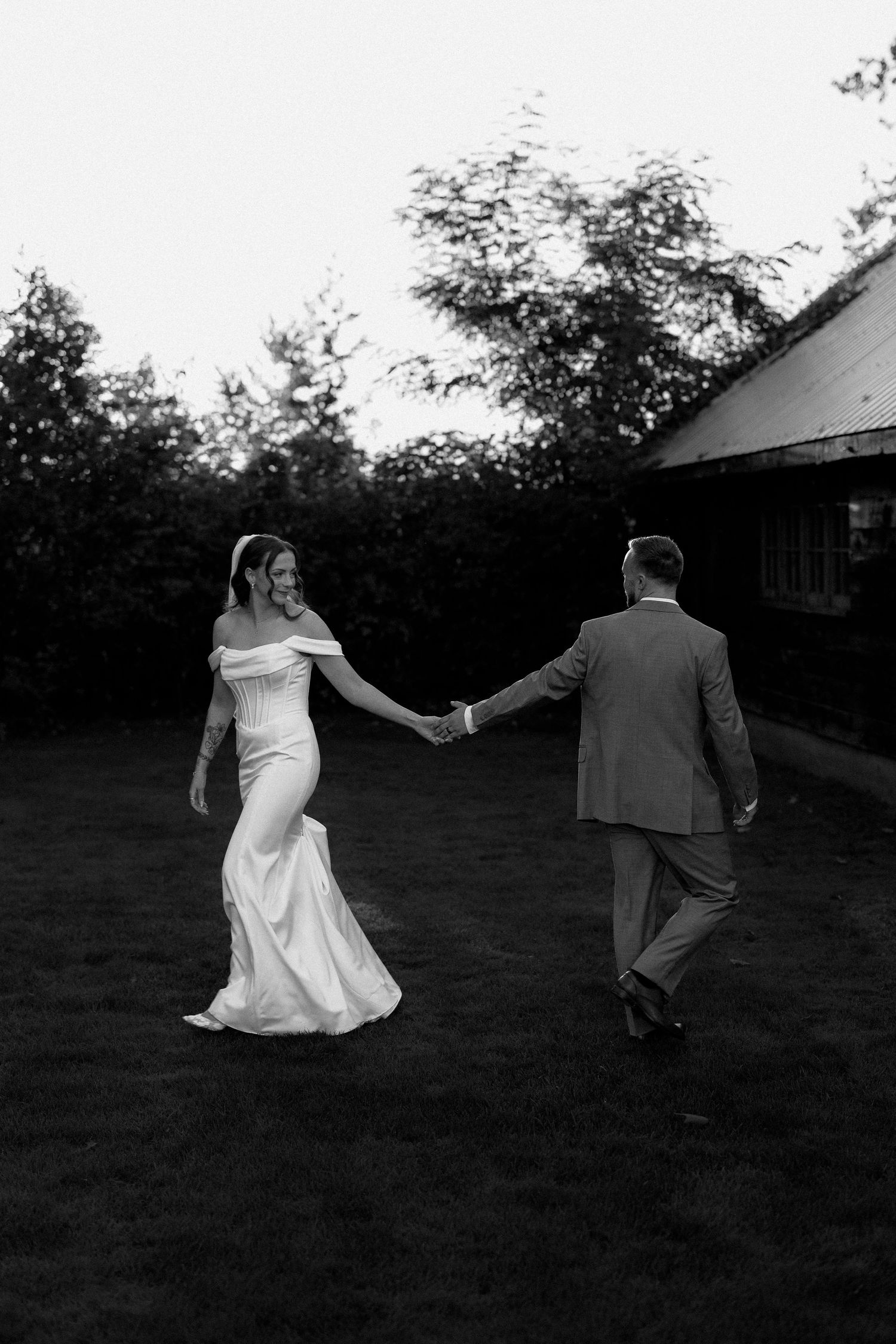 Black and white photo of a bride in a fitted gown holding hands with a groom in a suit outdoors.
