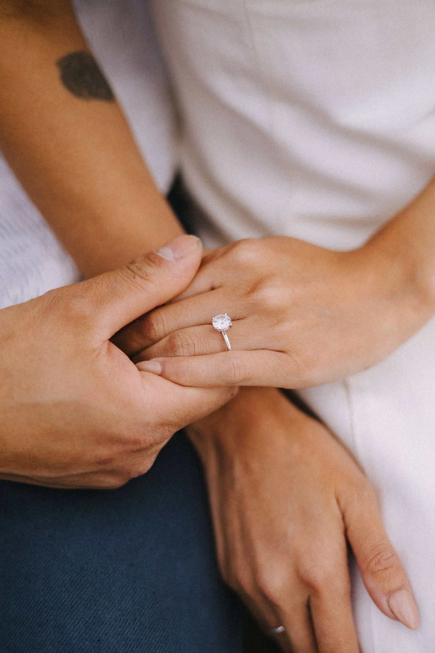 Close-up of hands wearing a delicate solitaire diamond engagement ring on a manicured finger against white and navy fabric.