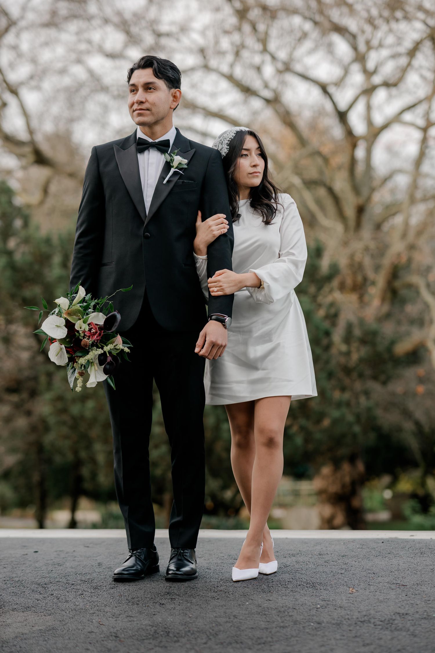 A couple in formal attire poses together on a path with trees in the background during a photo session.