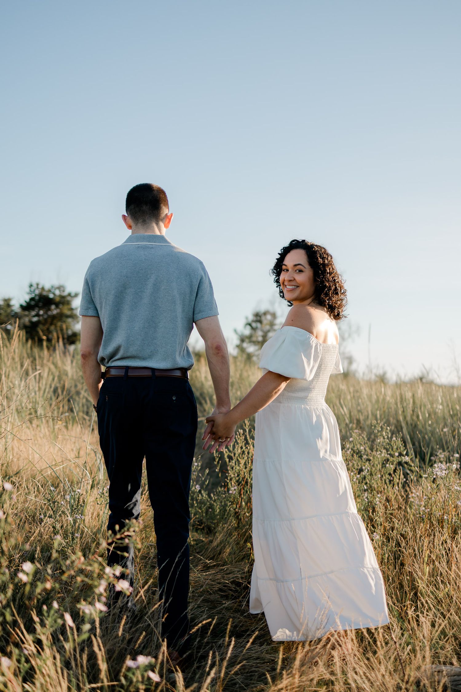A couple in formal attire walks hand in hand through a sunlit field of tall grass at golden hour.