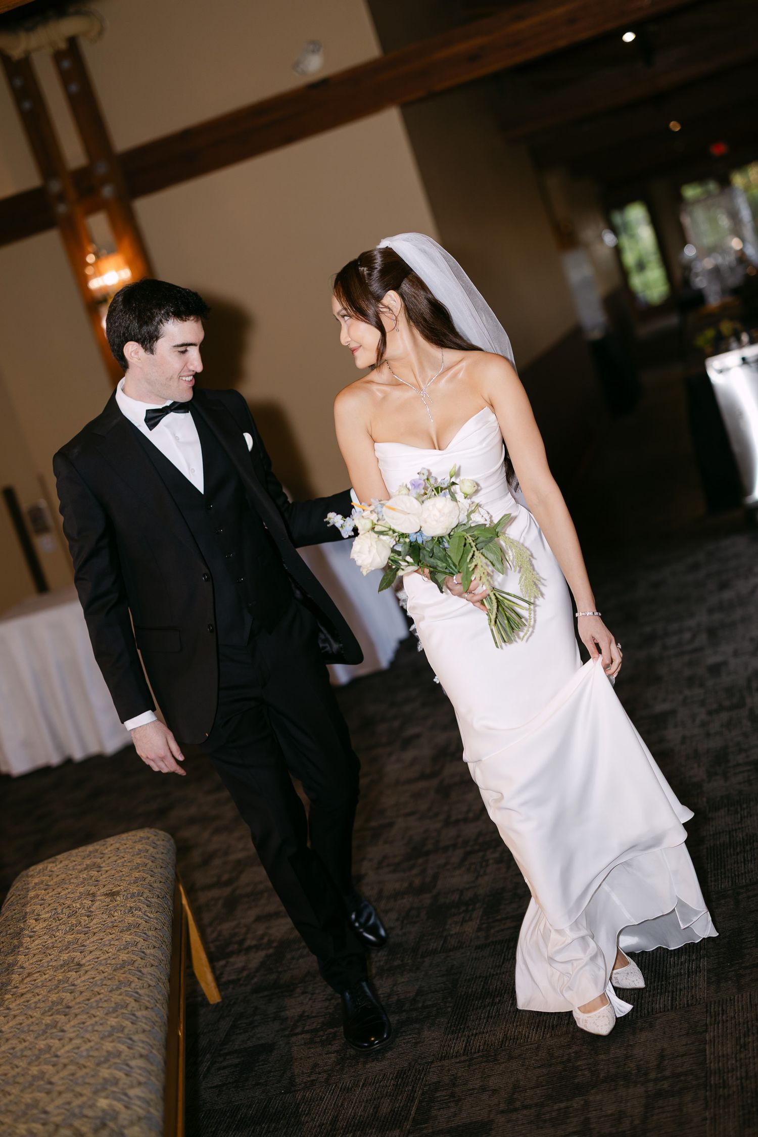 Bride in strapless gown holding white bouquet walks with groom in black tuxedo through wedding venue.