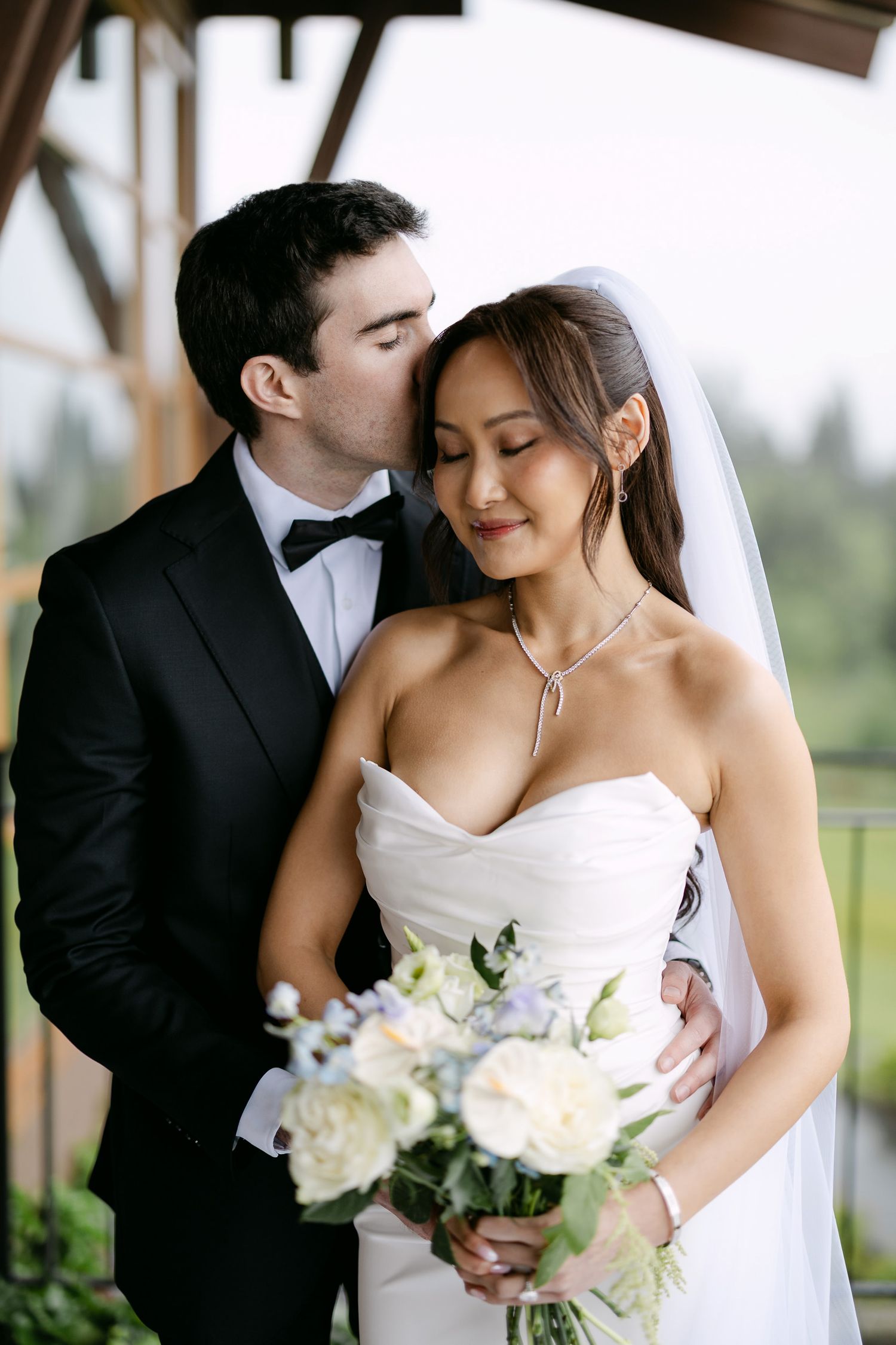 Groom in black tuxedo kissing bride on cheek as she smiles holding white and blue wedding bouquet.
