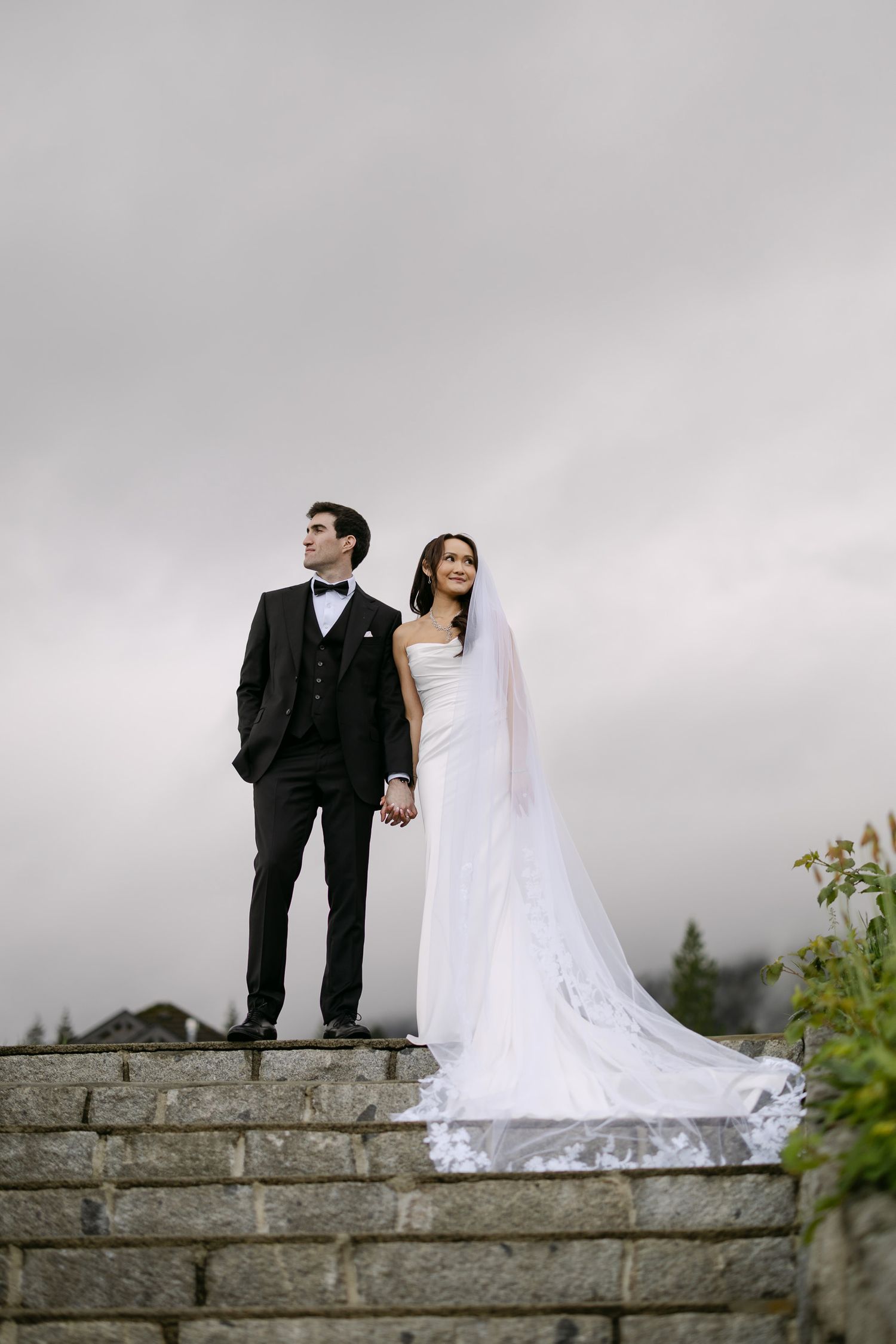 Bride and groom in formal wedding attire stand on stone steps holding hands under a cloudy sky.
