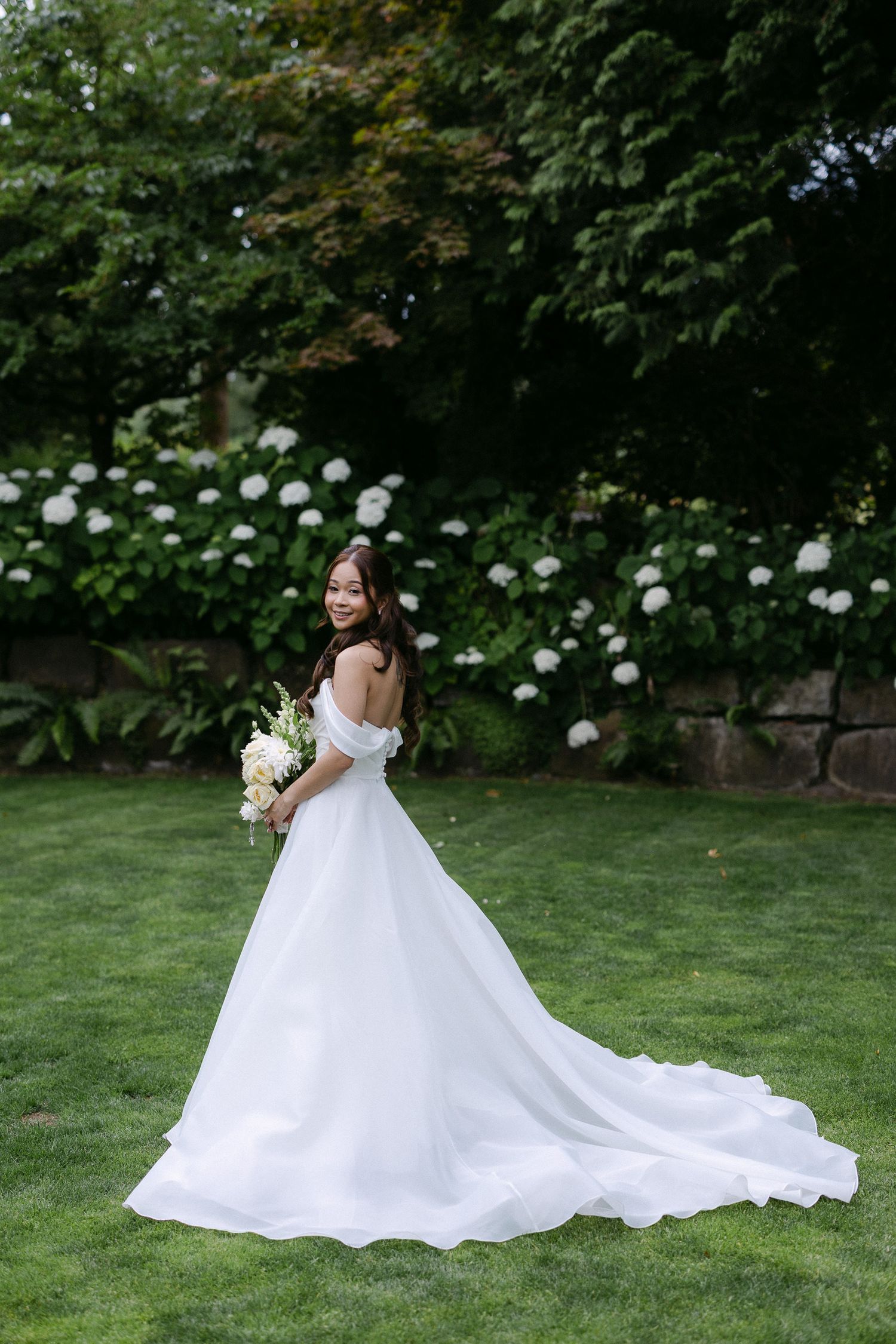 Bride in a white off-shoulder wedding gown smiling over her shoulder in a garden with hydrangeas.