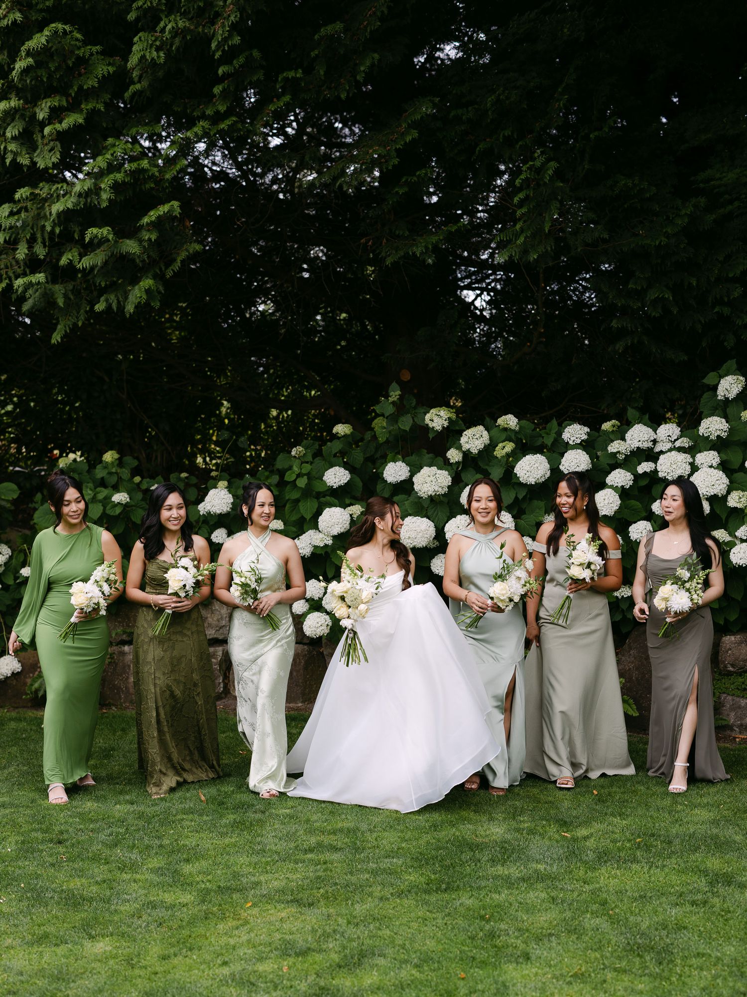 Bride in white gown with bridesmaids in green dresses holding bouquets outdoors near white hydrangeas.