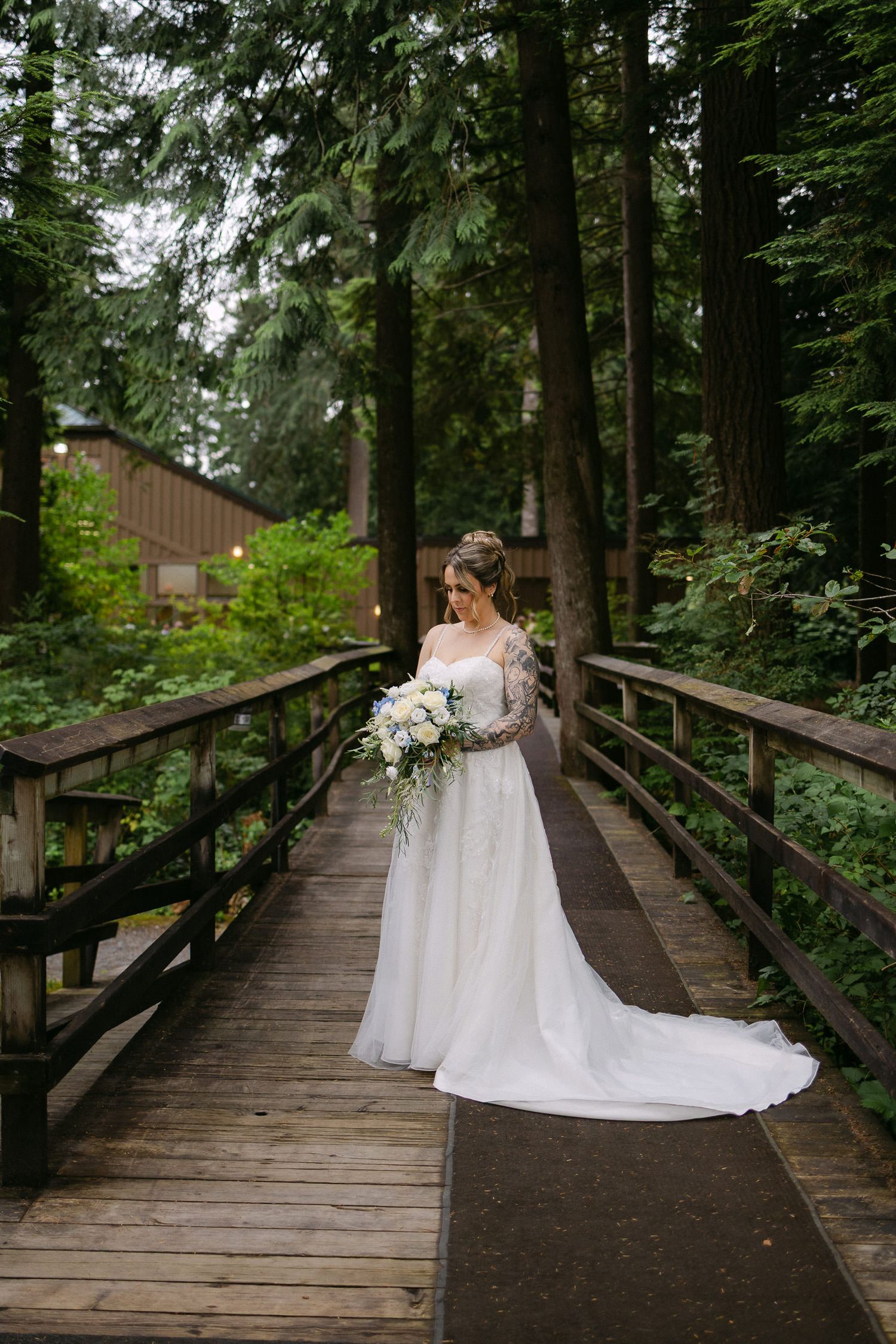 Bride in white lace gown holding bouquet stands on wooden bridge surrounded by tall trees in lush forest setting.