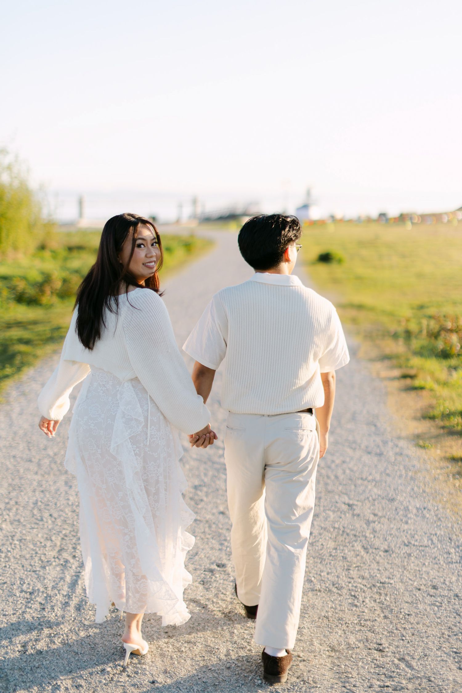 Smiling woman in a flowing white lace dress holds hands with a man in cream clothing as they walk down a gravel path at sunset.