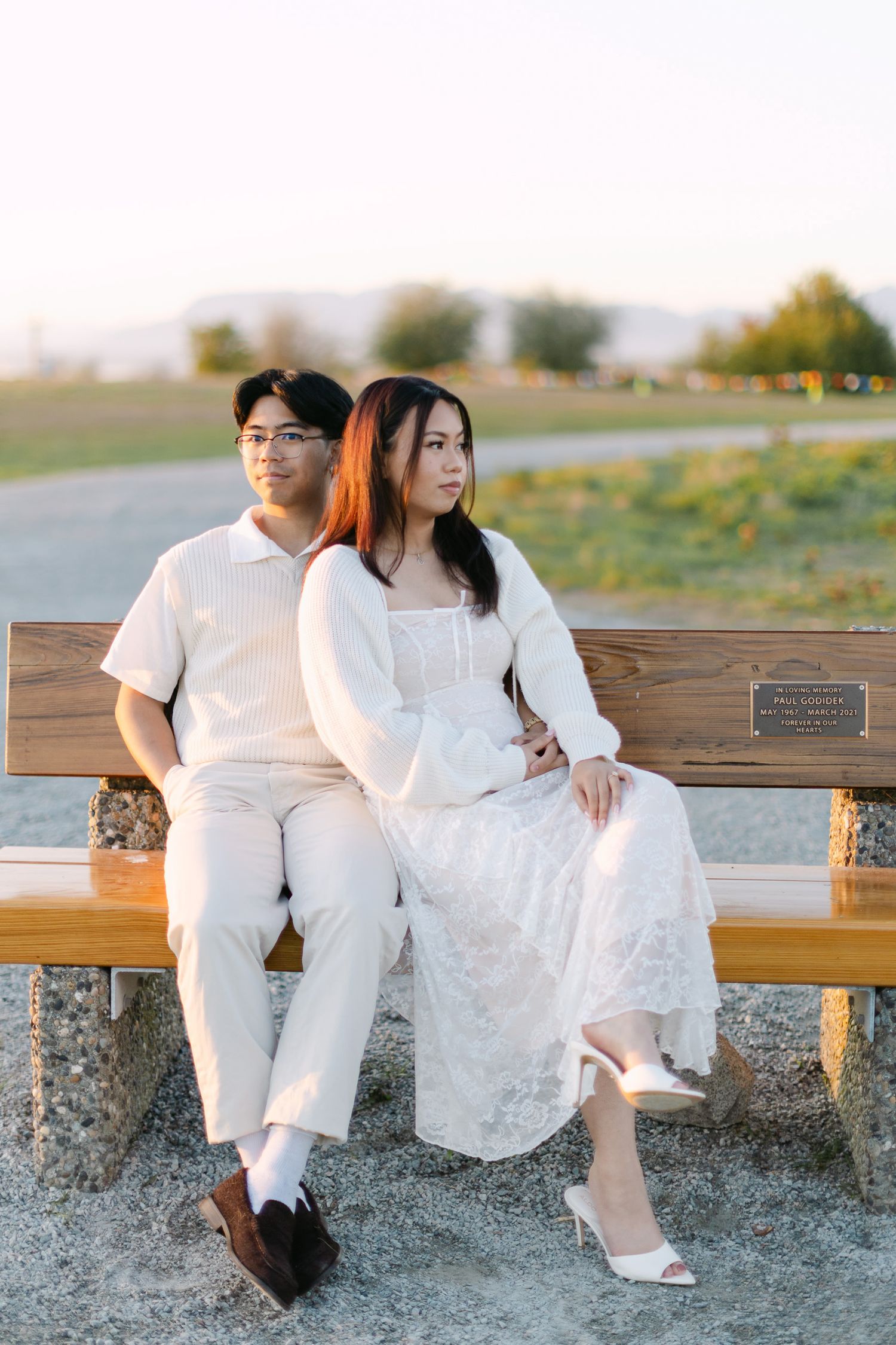 Couple in white outfits sitting together on a wooden bench in a grassy park during golden hour for an engagement photo.