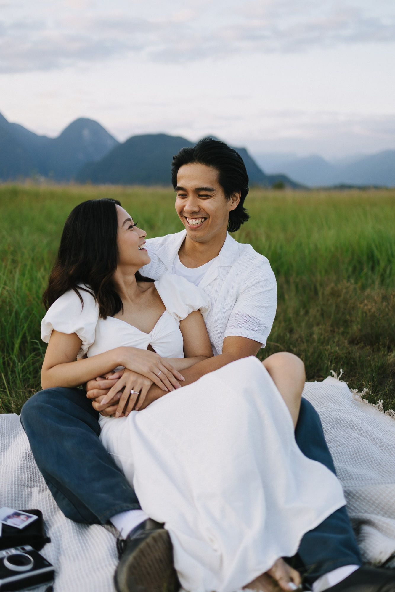 Smiling couple in white outfits sitting close together on a picnic blanket in a green grass field with mountains in the background.