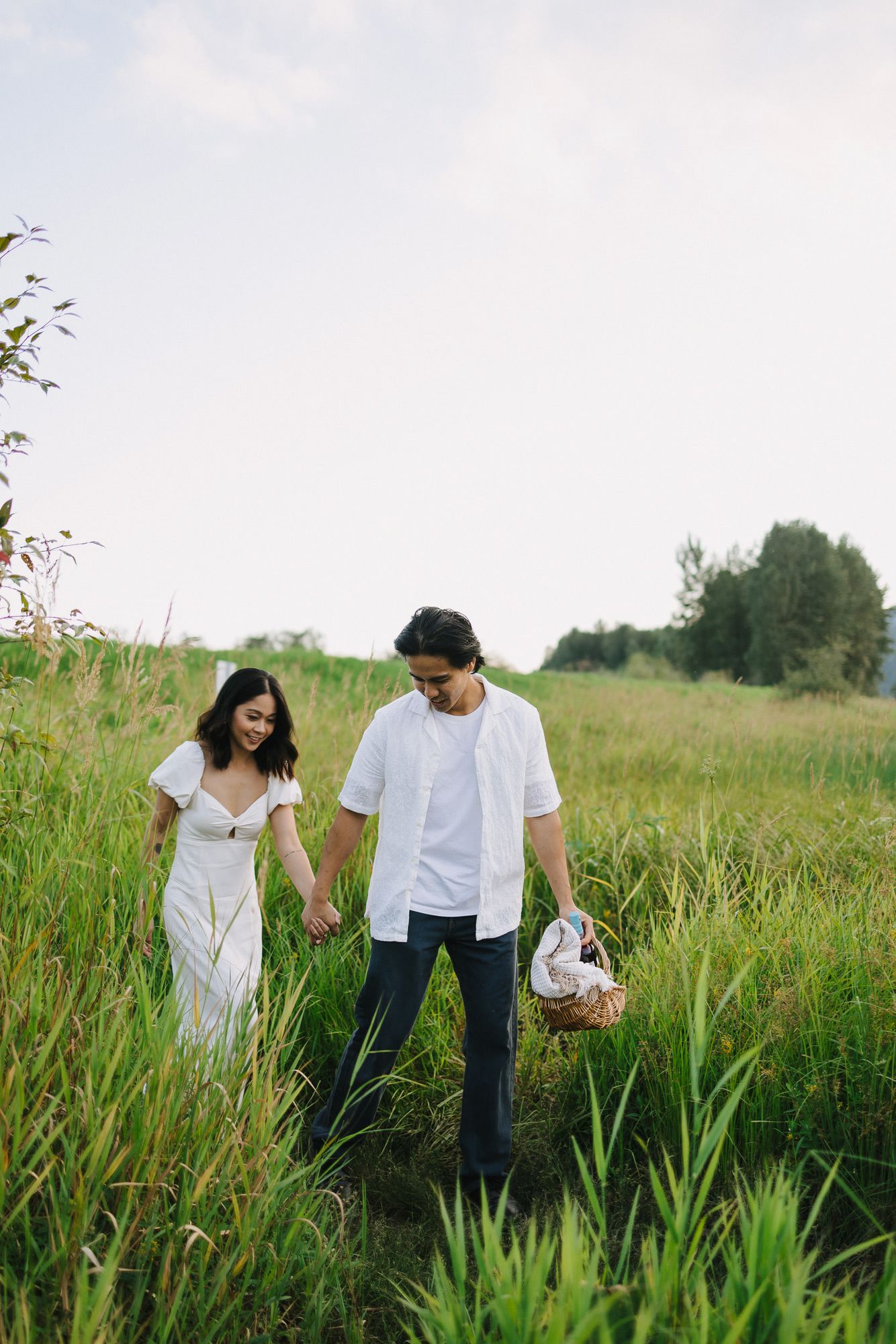 Smiling couple in white outfits holding hands and strolling through a green meadow with a wicker picnic basket.