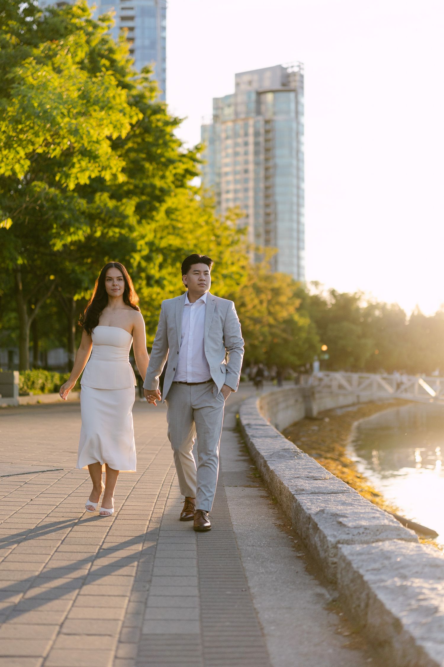 Couple holding hands while walking along a waterfront promenade at golden hour, with city high-rises and green trees in the background.