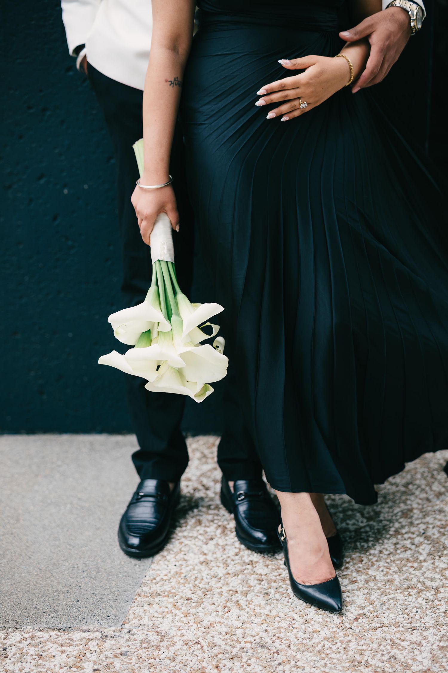 Close-up view of formal black shoes and dresses with white calla lily bouquets on gravel ground at an elegant event.