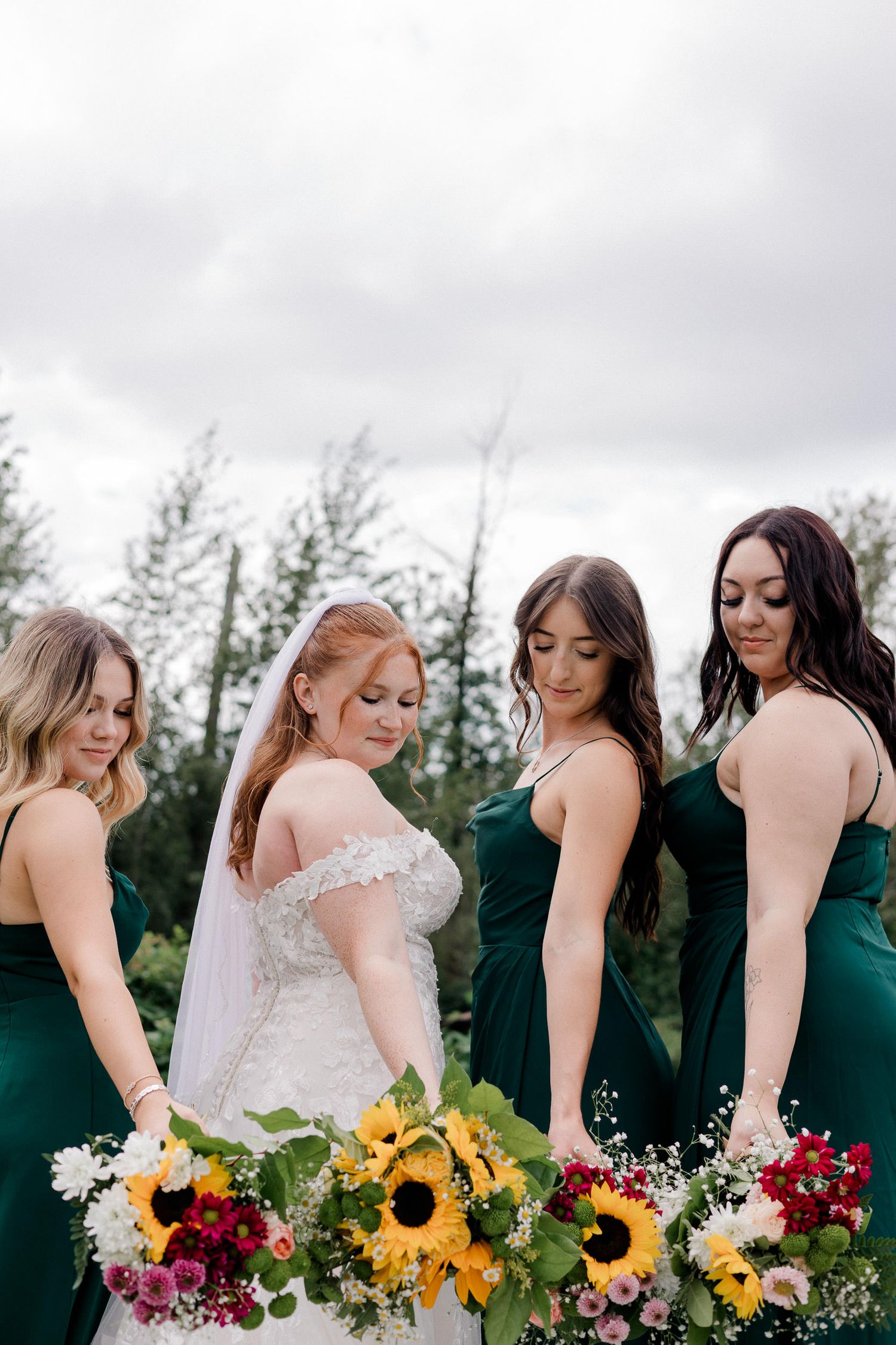 A line of bridesmaids in dark green dresses holding colorful bouquets with sunflowers and red flowers pose together outdoors.