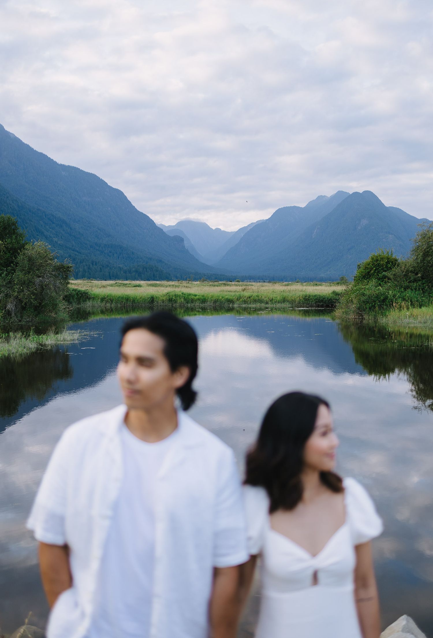 Couple in white outfits standing by a scenic mountain lake with reflective waters and misty peaks in the background.