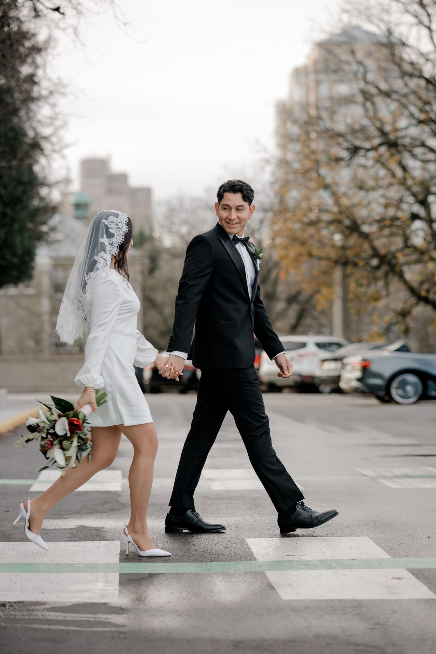 Couple in wedding attire playfully walking across crosswalk in modern urban setting.