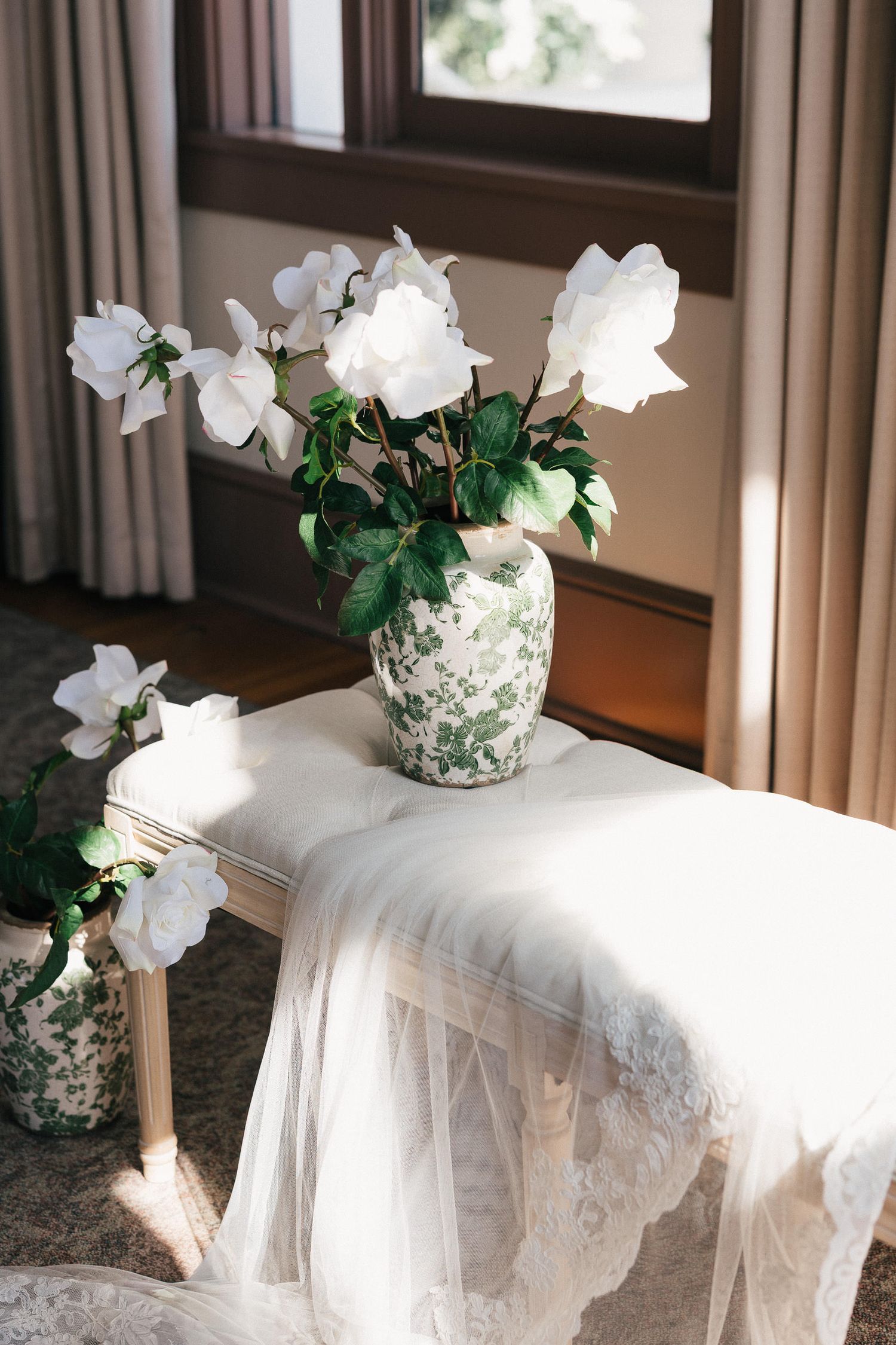 A delicate white wedding veil is draped across chairs with white floral arrangements in vintage vases on display.