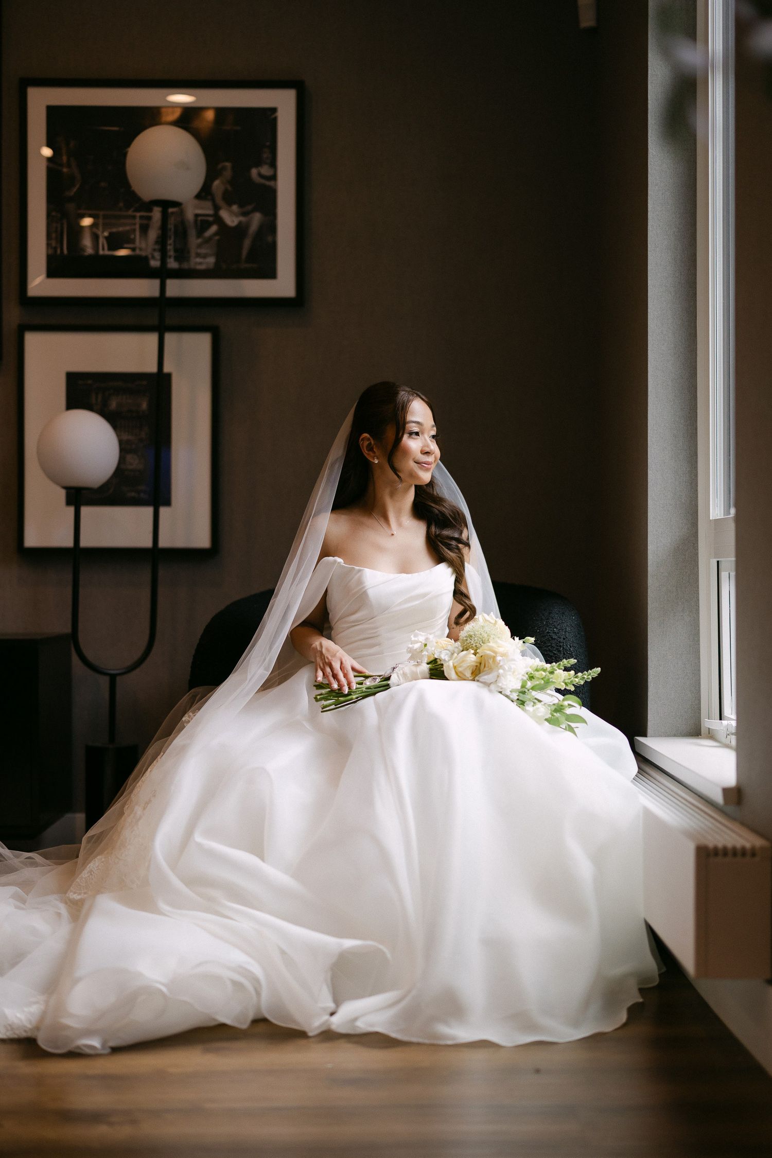 Bride in strapless white ball gown with veil holds white bouquet, seated by window in elegant dark interior.