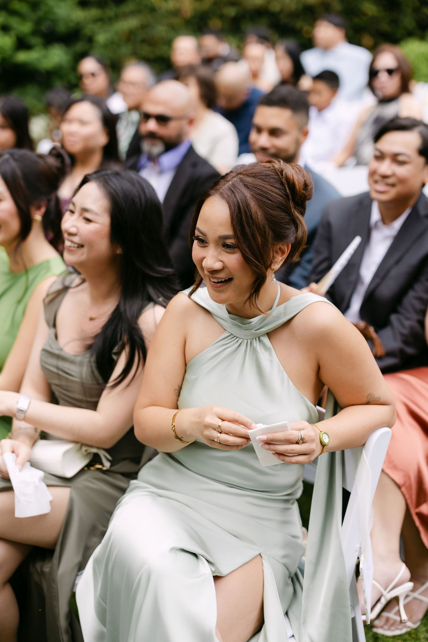 Bridesmaid in sage green halter dress laughing joyfully at outdoor wedding ceremony with guests seated behind her.