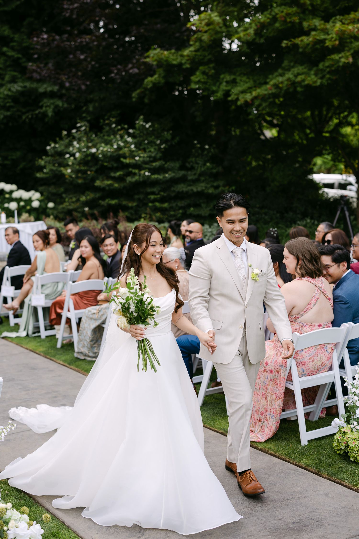 Newlywed couple walks down outdoor aisle, bride in white gown holding bouquet, groom in beige suit, guests seated on white chairs.