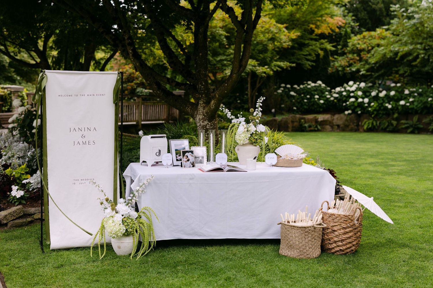 Elegant outdoor wedding welcome table with white linen, floral arrangements, and signage on green lawn.