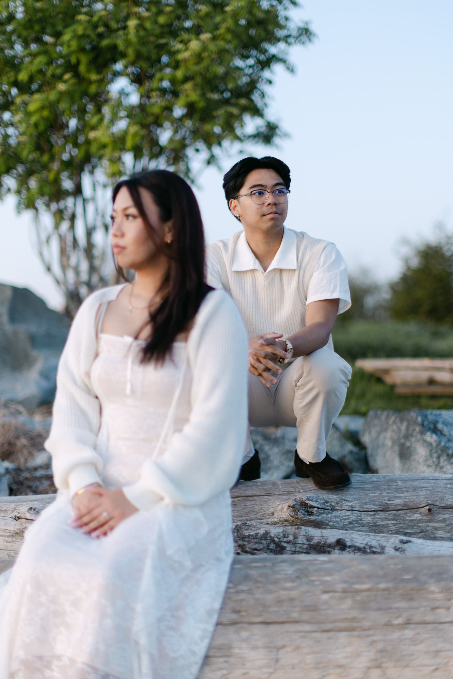 Couple in white outfits sit on a driftwood log at a beach during golden hour, with the woman in a lace dress in the foreground.