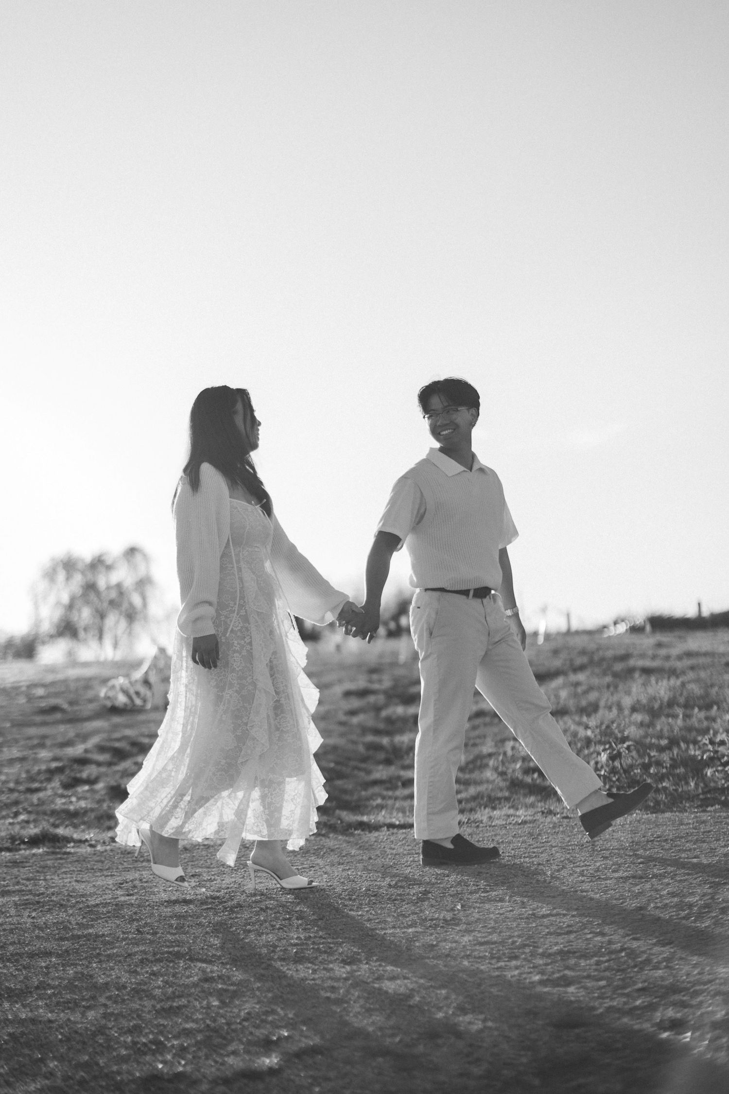 Black and white photo of a couple holding hands and walking across a grassy field, she in a lace dress and he in a polo and cap.