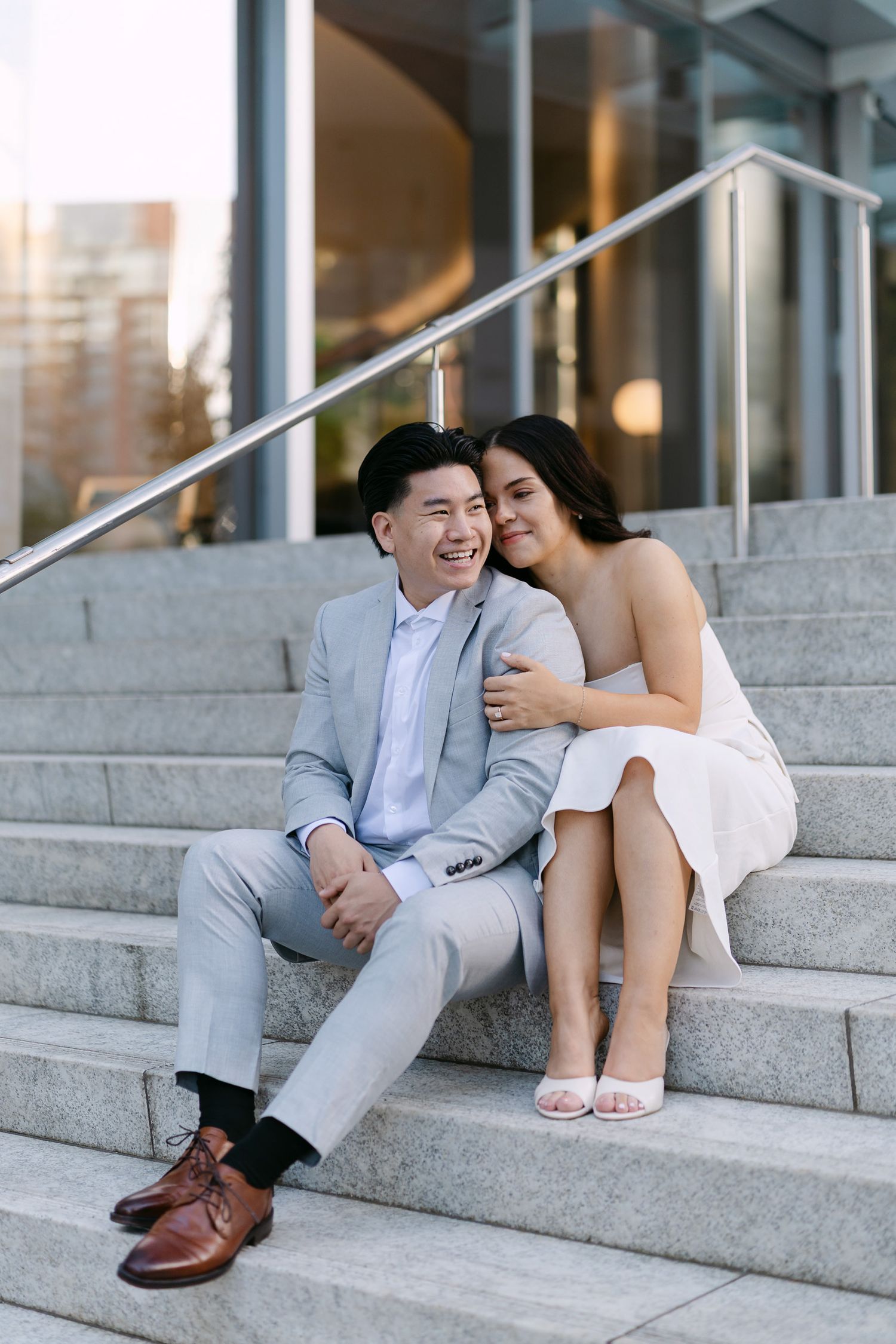 A laughing man in a gray suit sits on stone steps while a woman in a white dress leans affectionately against his shoulder.