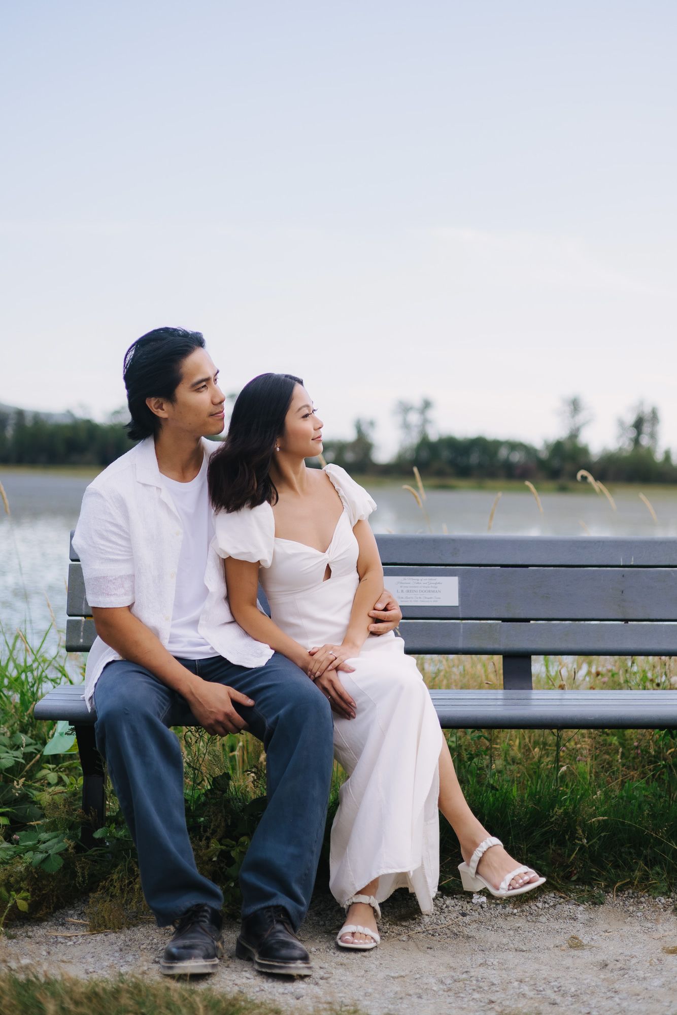 Couple in casual white outfits sitting close together on a lakeside bench during an engagement photo session.